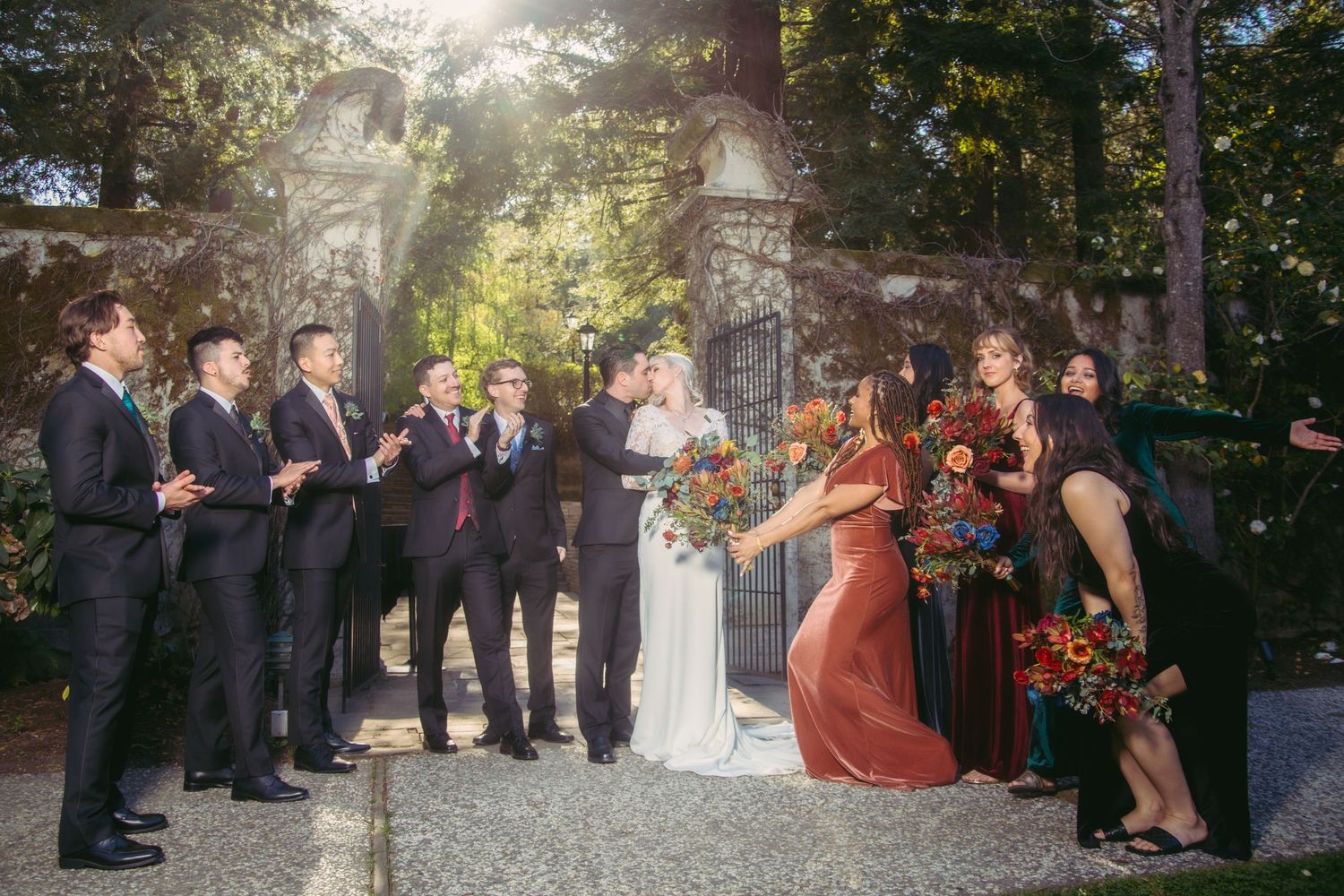 Wedding party celebrating at ornate garden gates, bridesmaids in jewel-tone velvet gowns with colorful bouquets.