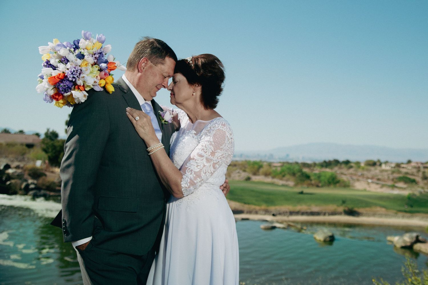 Bride and groom sharing a romantic moment outdoors near a river, bride holding a colorful bouquet, mountains in the background.