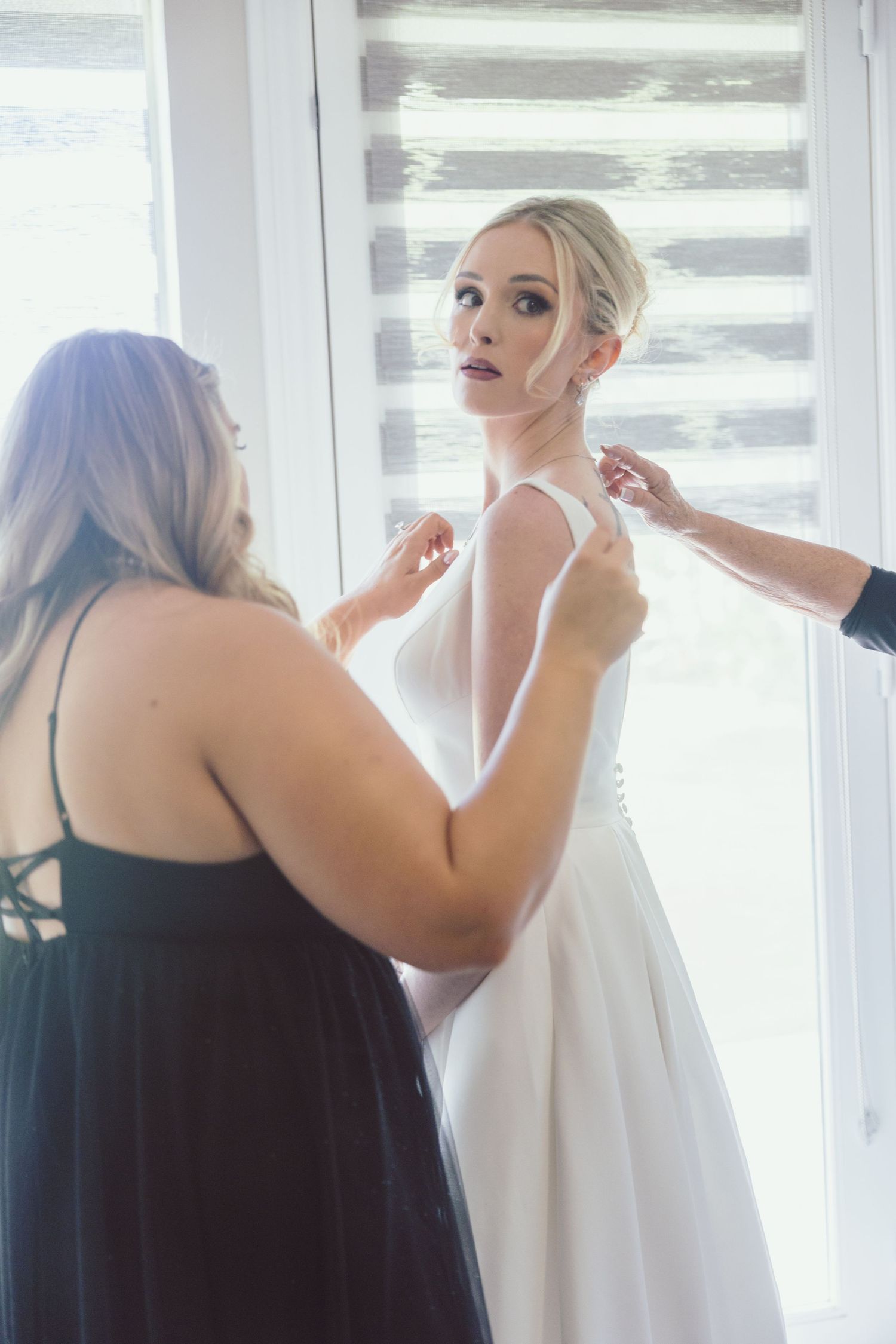 Bride in white wedding gown being helped by bridesmaids near bright window.