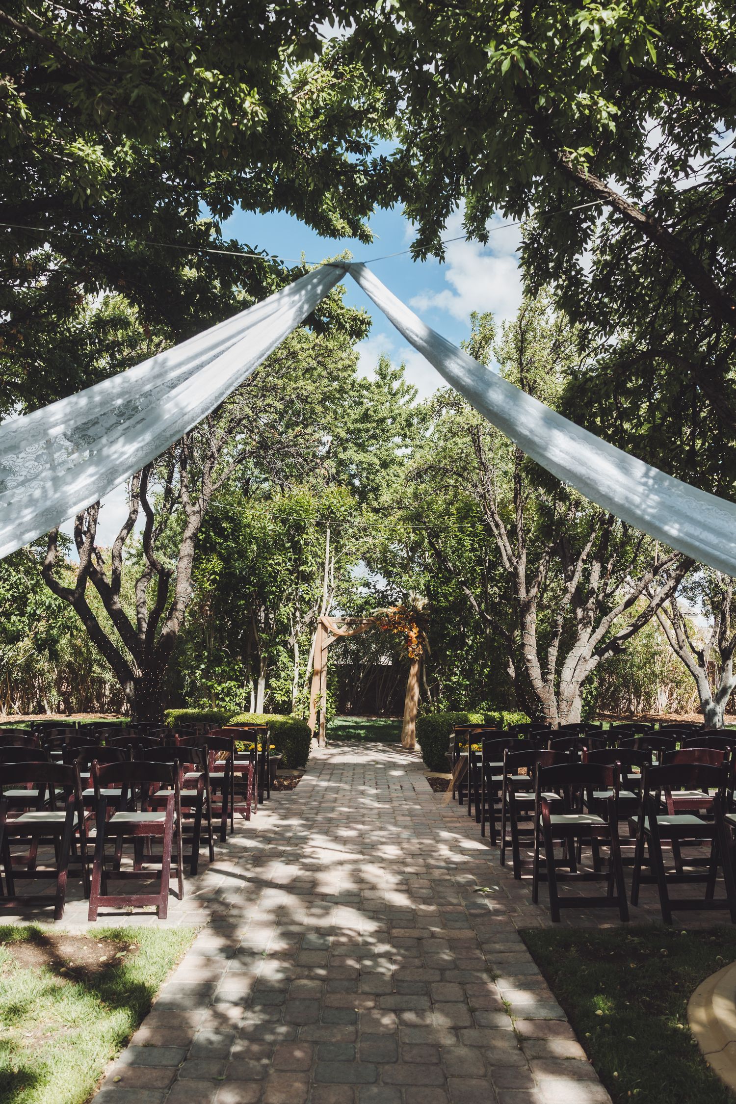 Outdoor wedding ceremony aisle lined with wooden chairs under white draping and lush green trees.