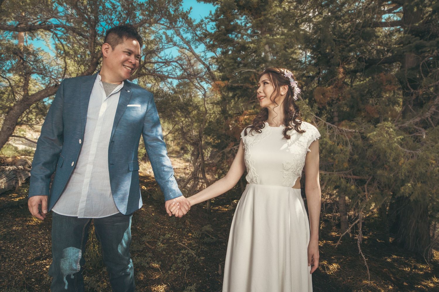 A couple in wedding attire holds hands outdoors among pine trees, the groom in blue suit and bride in white dress.