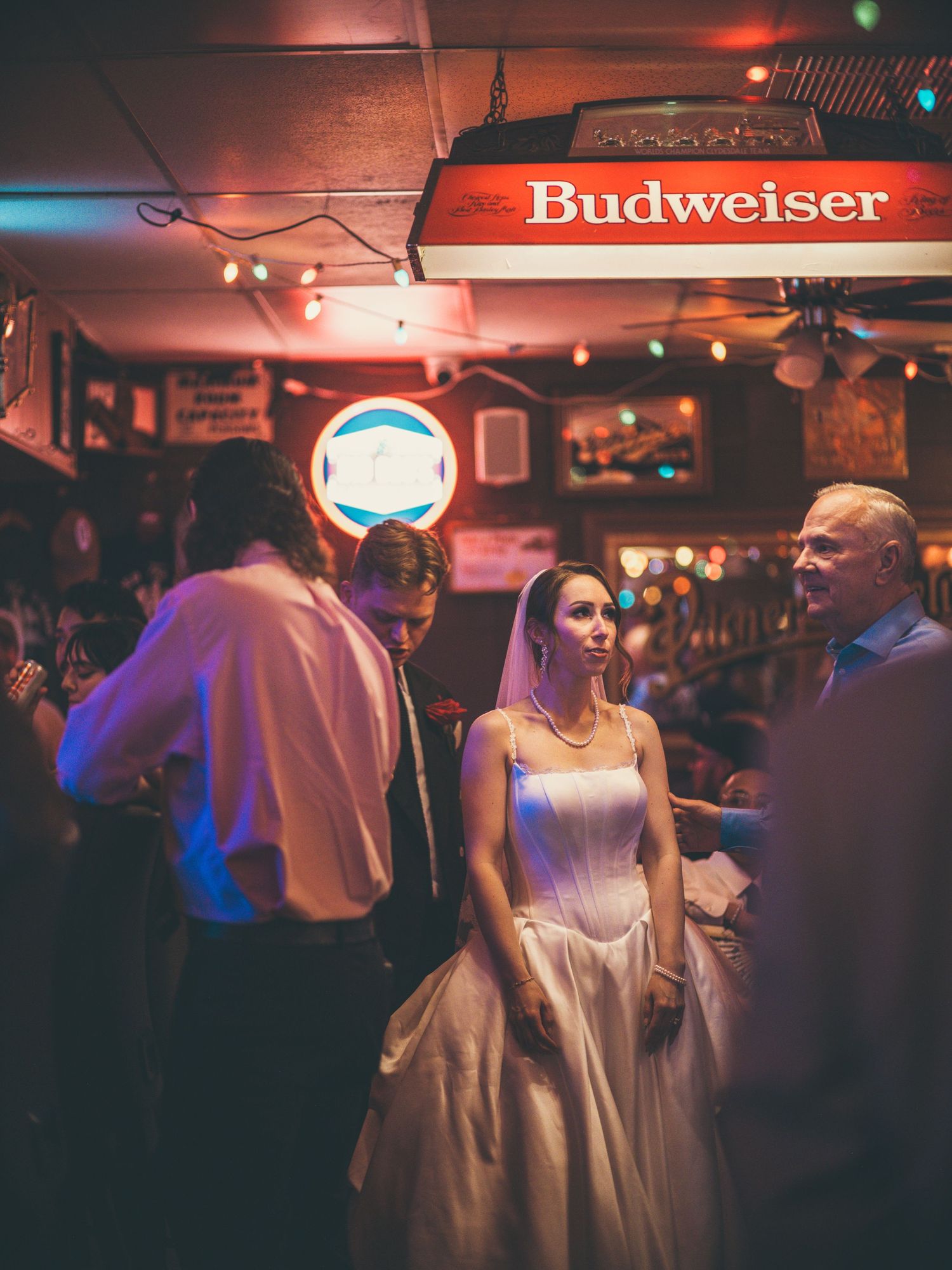 Bride in white wedding gown and veil socializing at a bar venue under a Budweiser sign during reception.