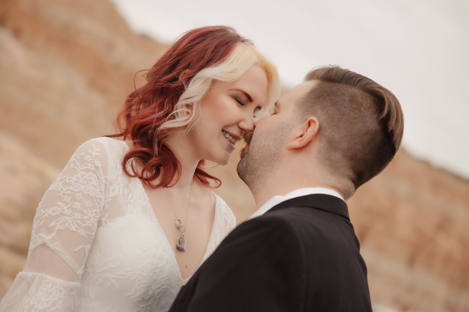 Smiling bride with red hair in lace wedding dress shares romantic moment with groom outdoors.