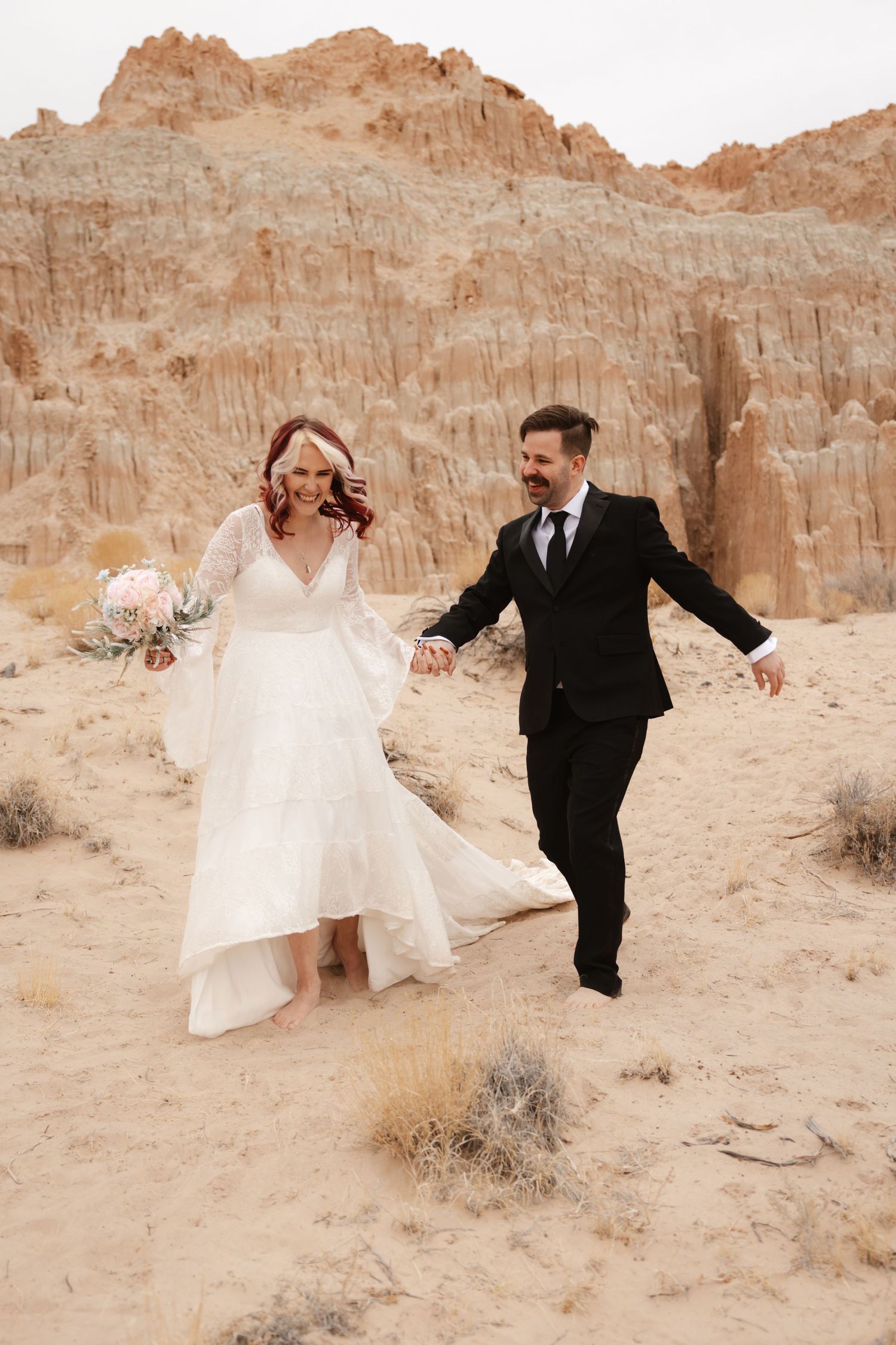 Joyful bride and groom run hand in hand through sandy desert terrain with rocky cliffs behind them.