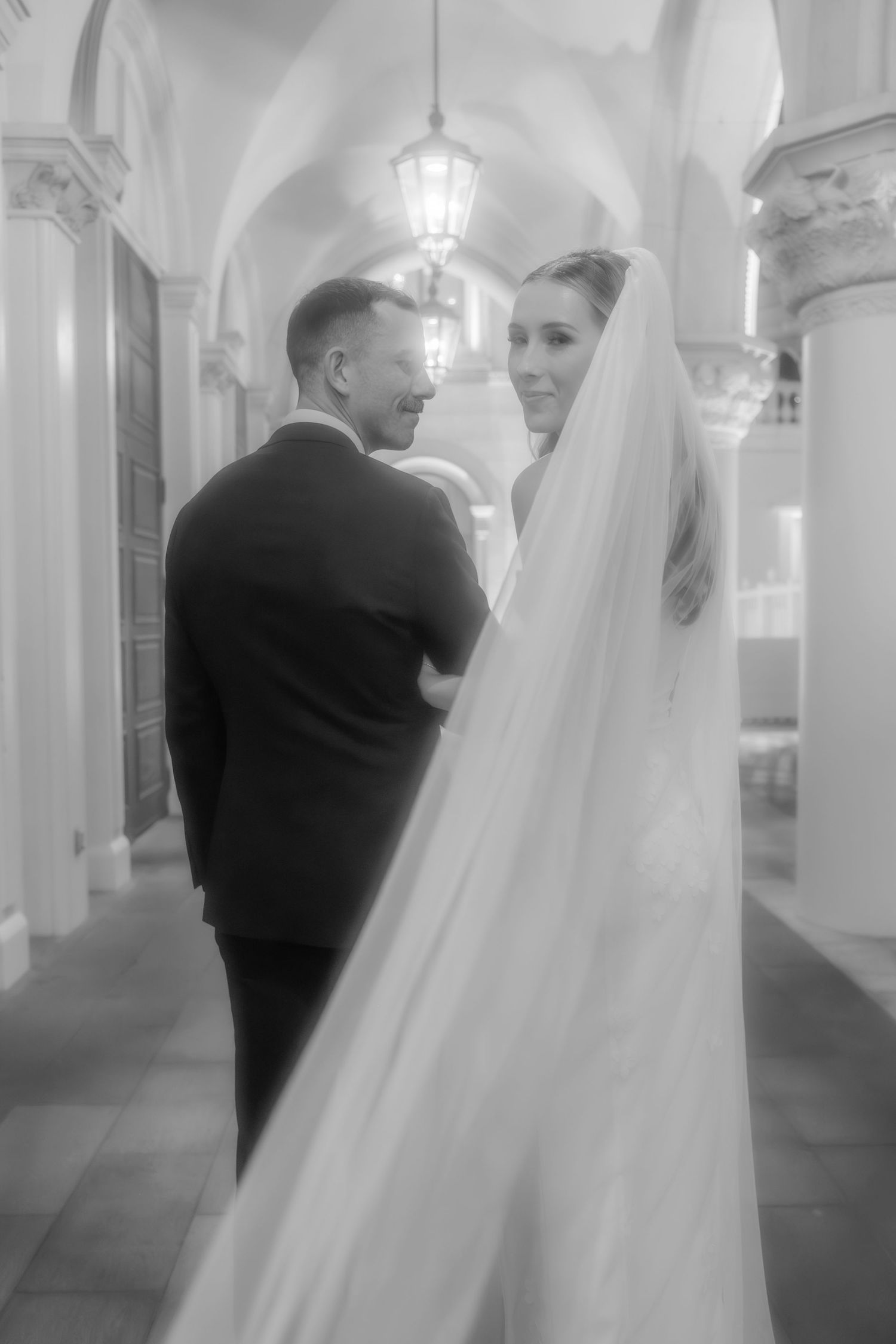 Black and white portrait of bride and groom in arched hallway with flowing veil.