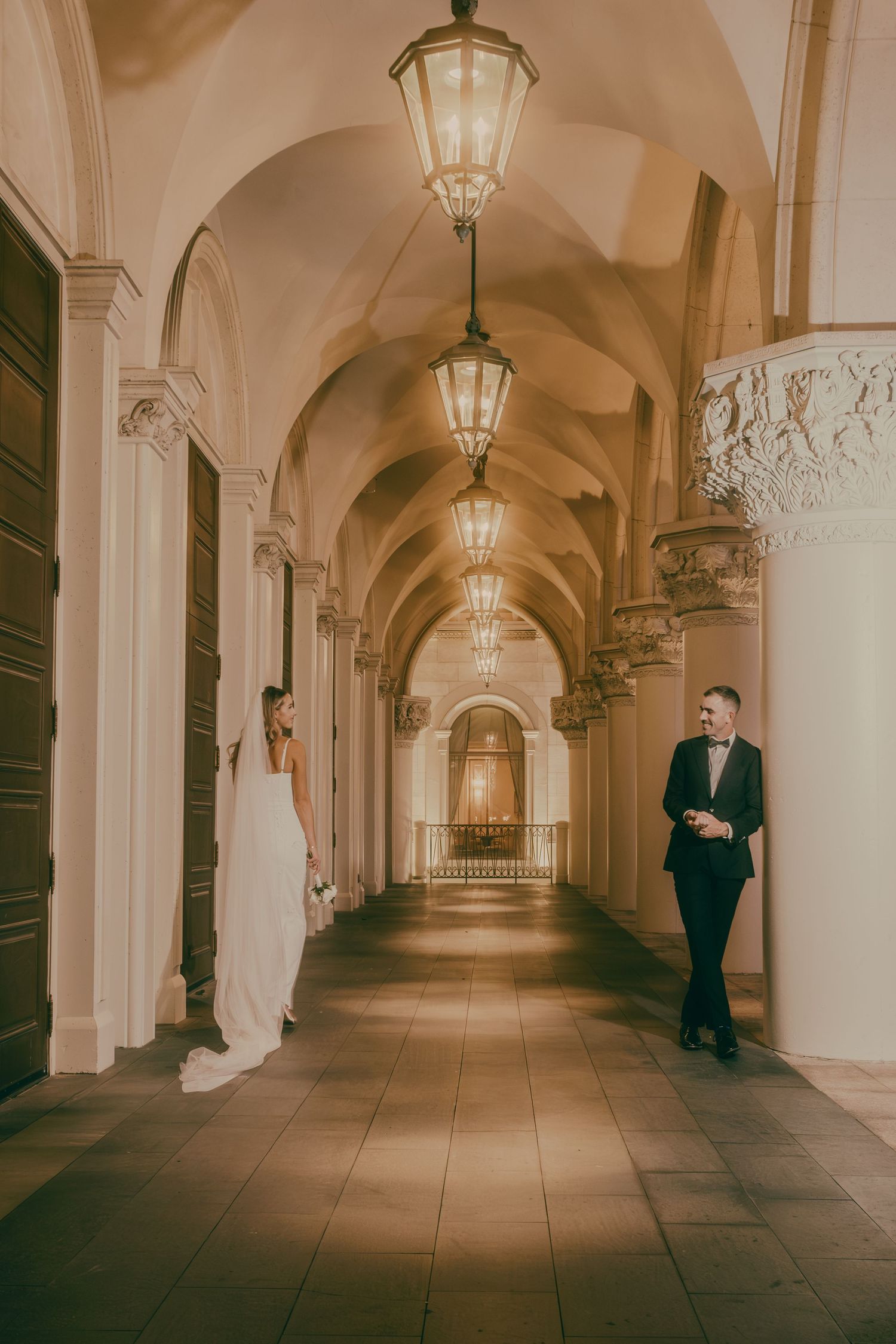 Bride in white gown and groom in tuxedo stand apart in dramatic arched corridor with lanterns.