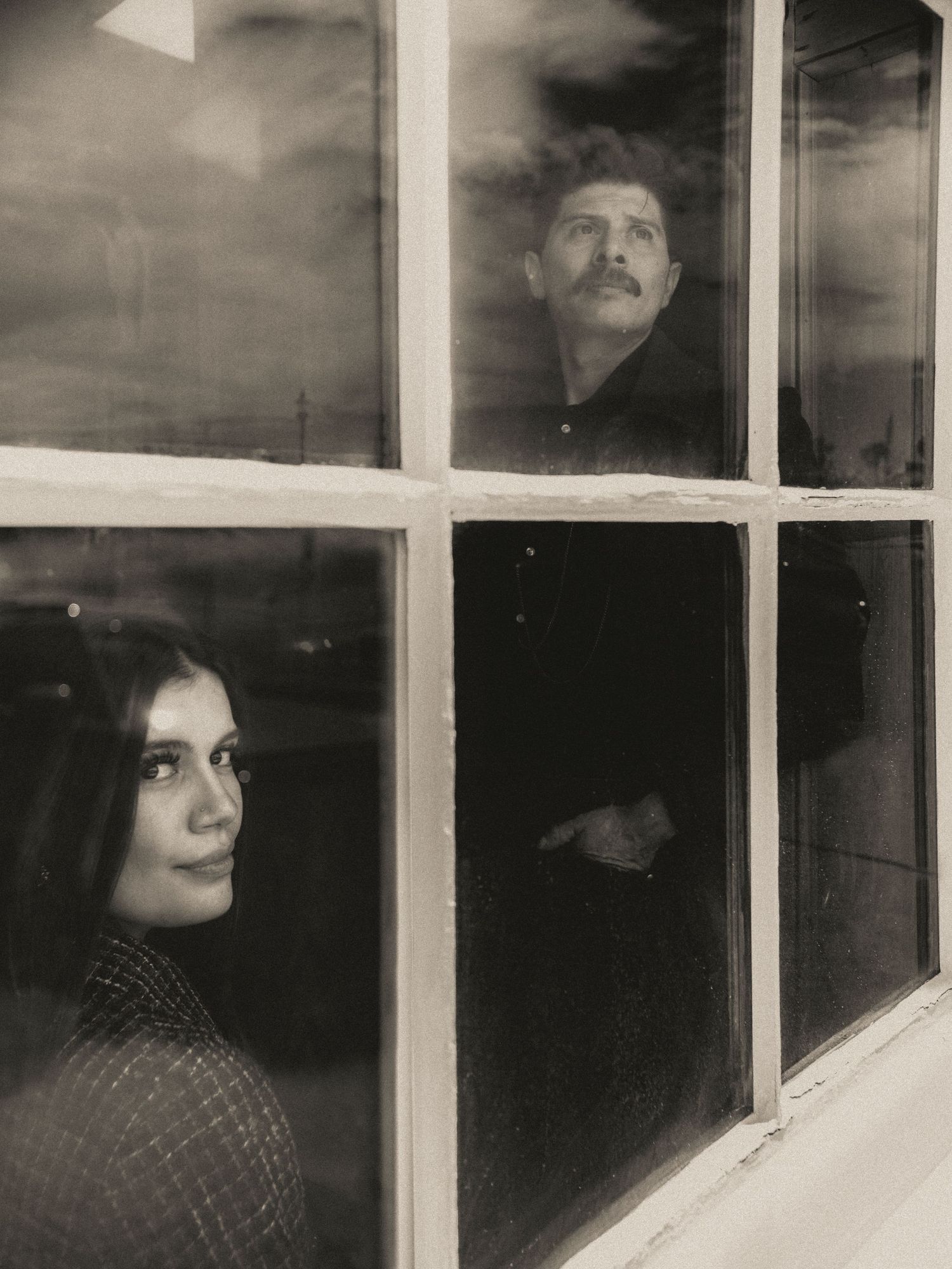 Black and white photo of a man with mustache and woman gazing through a vintage window pane.