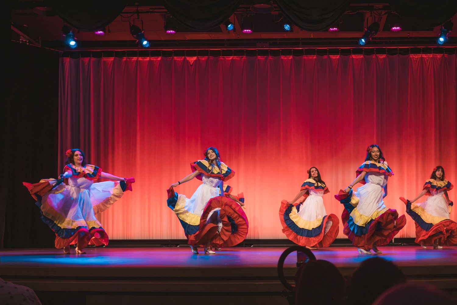 Colorful folkloric dancers perform on a red-curtained stage in vibrant traditional dresses.