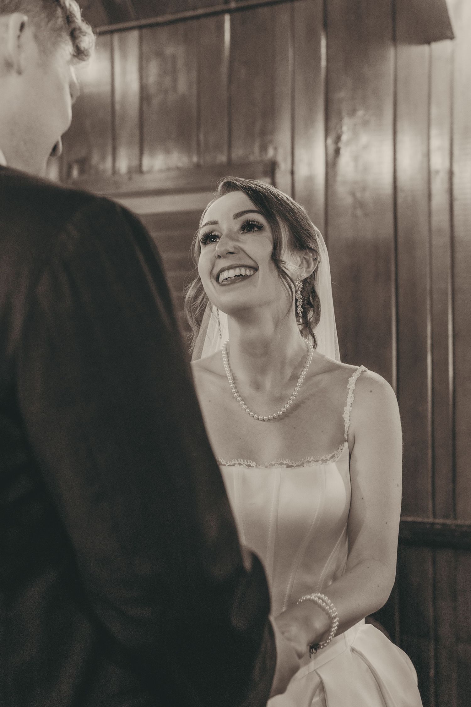 Black and white photo of a smiling bride in a pearl necklace and veil holding hands with groom during wedding ceremony.