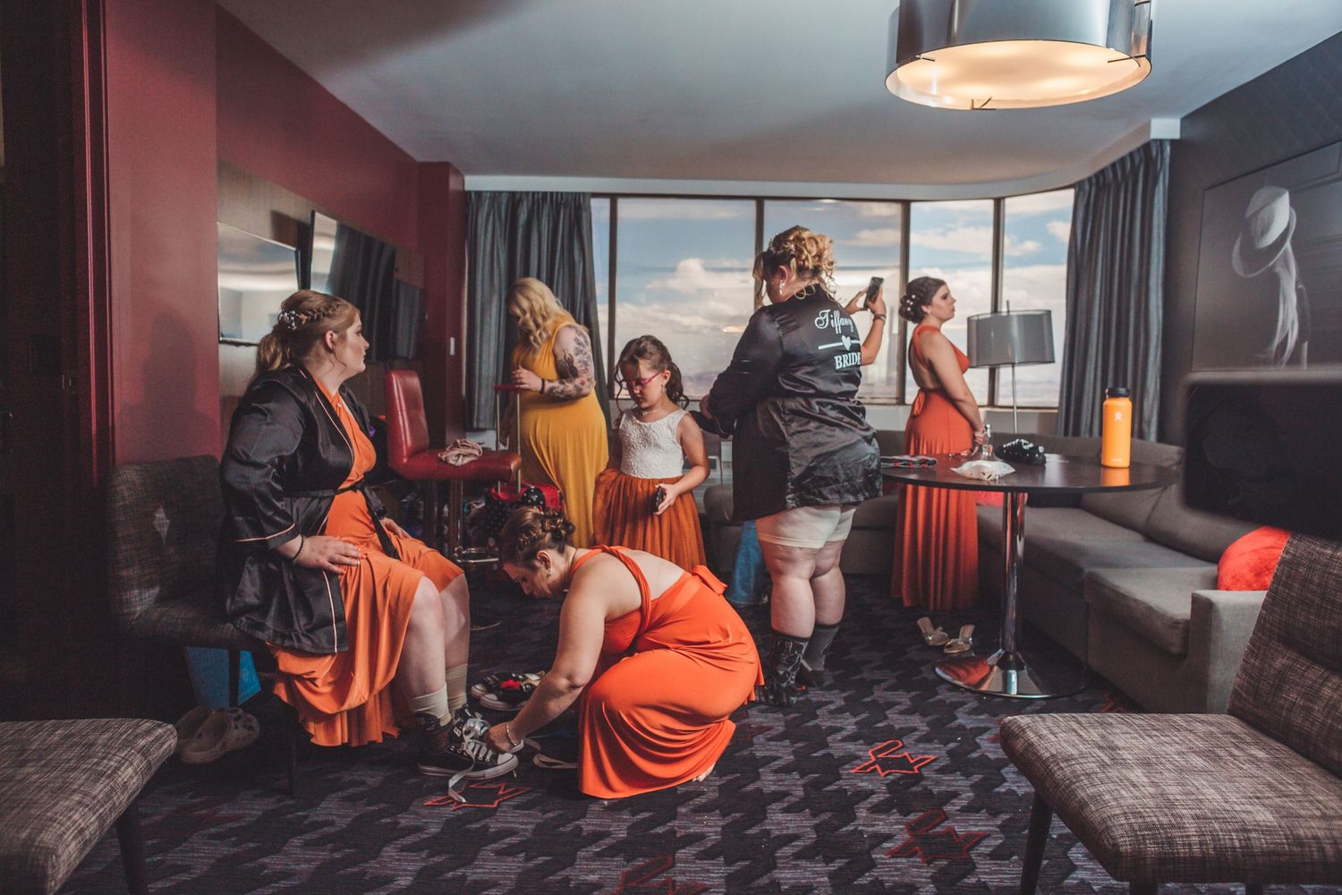 Women in orange dresses prepare together in a hotel room, helping each other get ready for a special event.