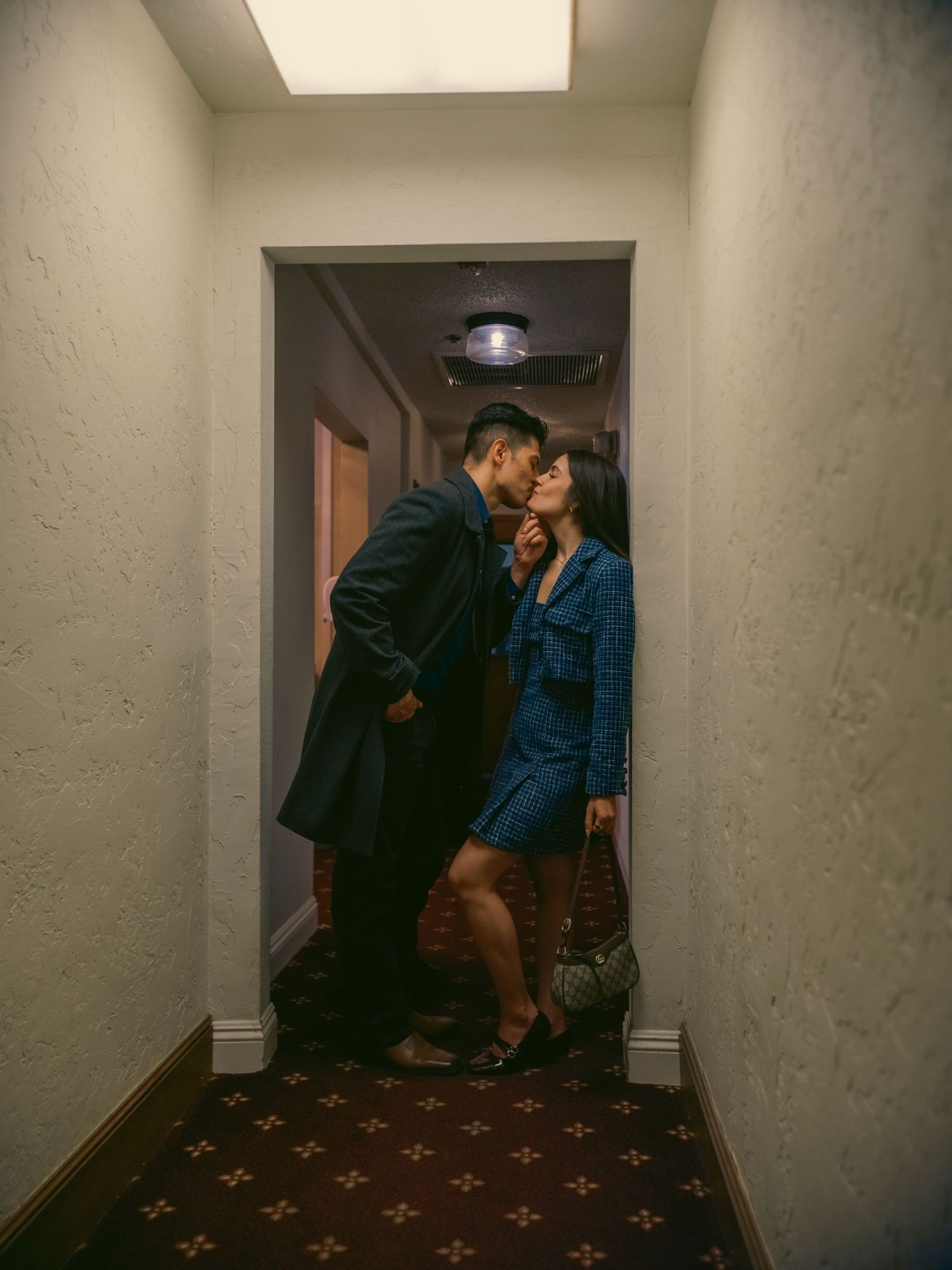 Couple sharing a romantic kiss in a hotel hallway, man in navy coat and woman in blue denim dress.