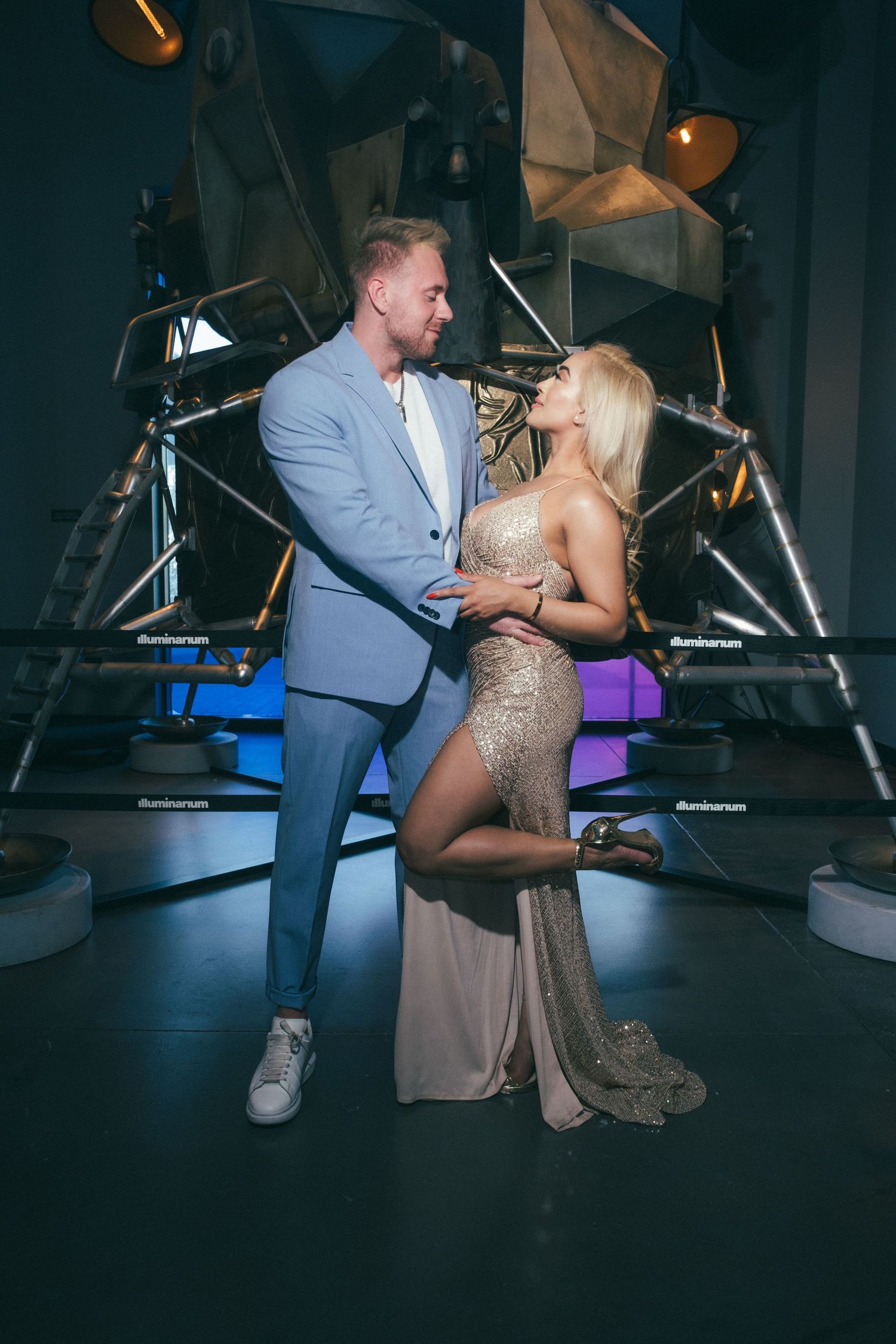 Elegant couple posing together in a glamorous venue, man in blue suit and woman in gold sequin gown.