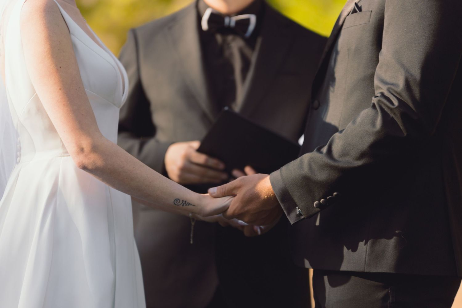 Bride and groom holding hands during wedding ceremony with officiant in background.