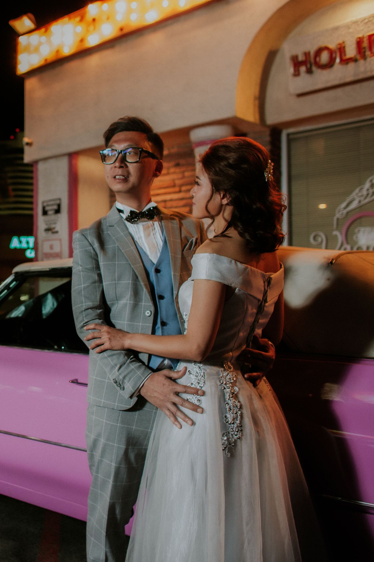 A couple in wedding attire poses by a pink car under neon lights, the groom in a plaid suit and the bride in a white gown.