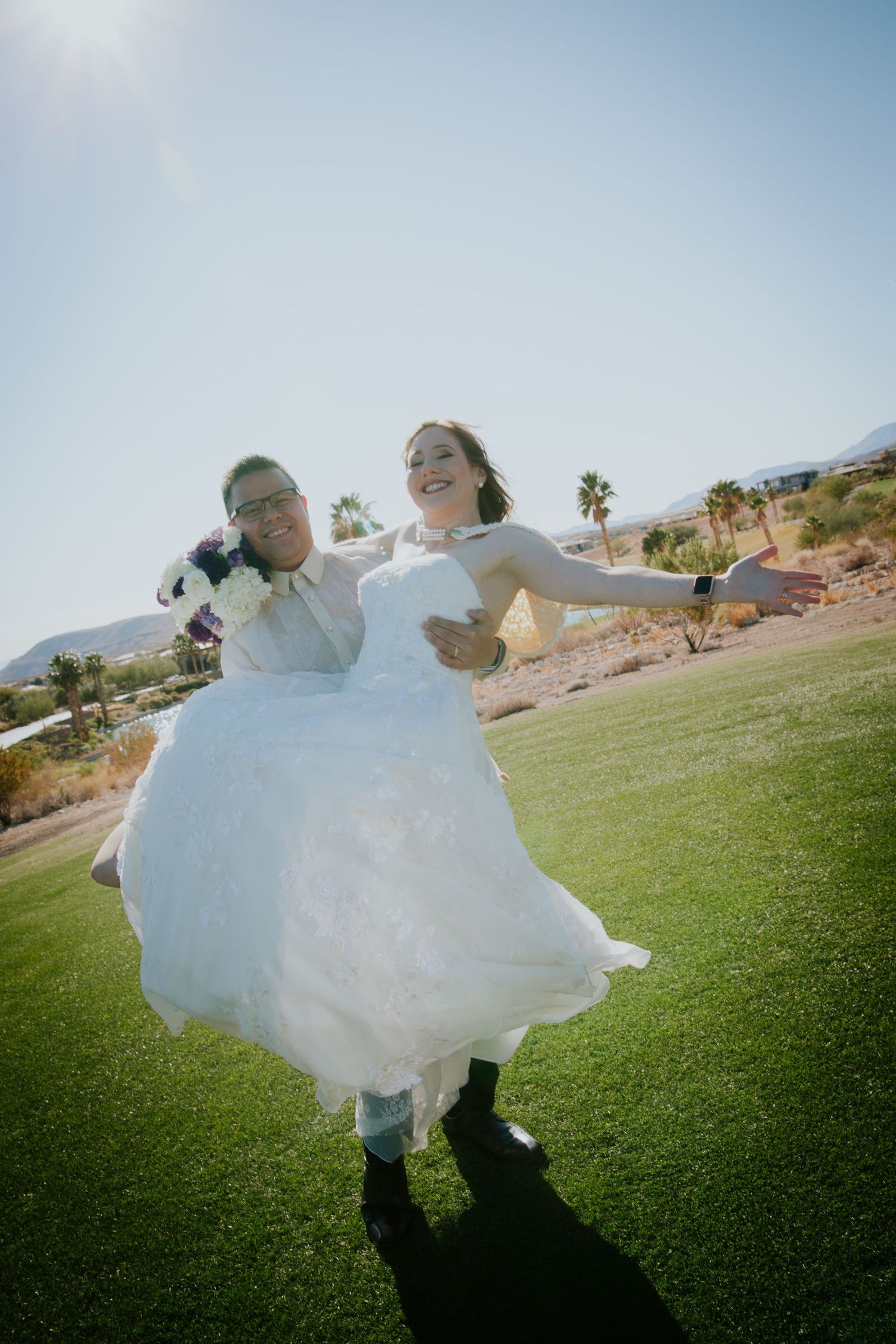 Two brides celebrate joyfully outdoors on green grass, one lifting the other in a white wedding gown, with palm trees and mountains.