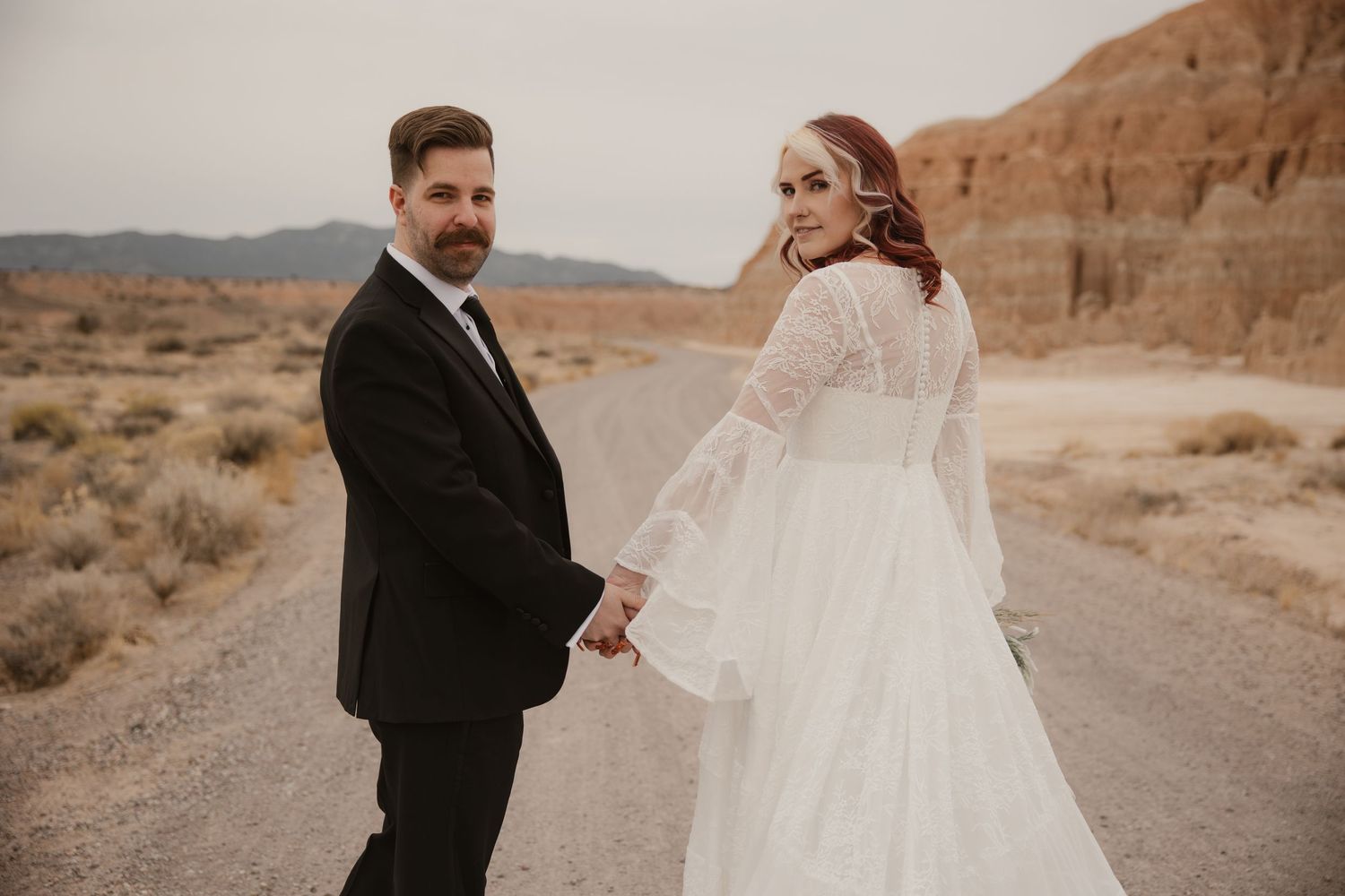 Bride in lace gown and groom hold hands on desert road with red rock formations in background.