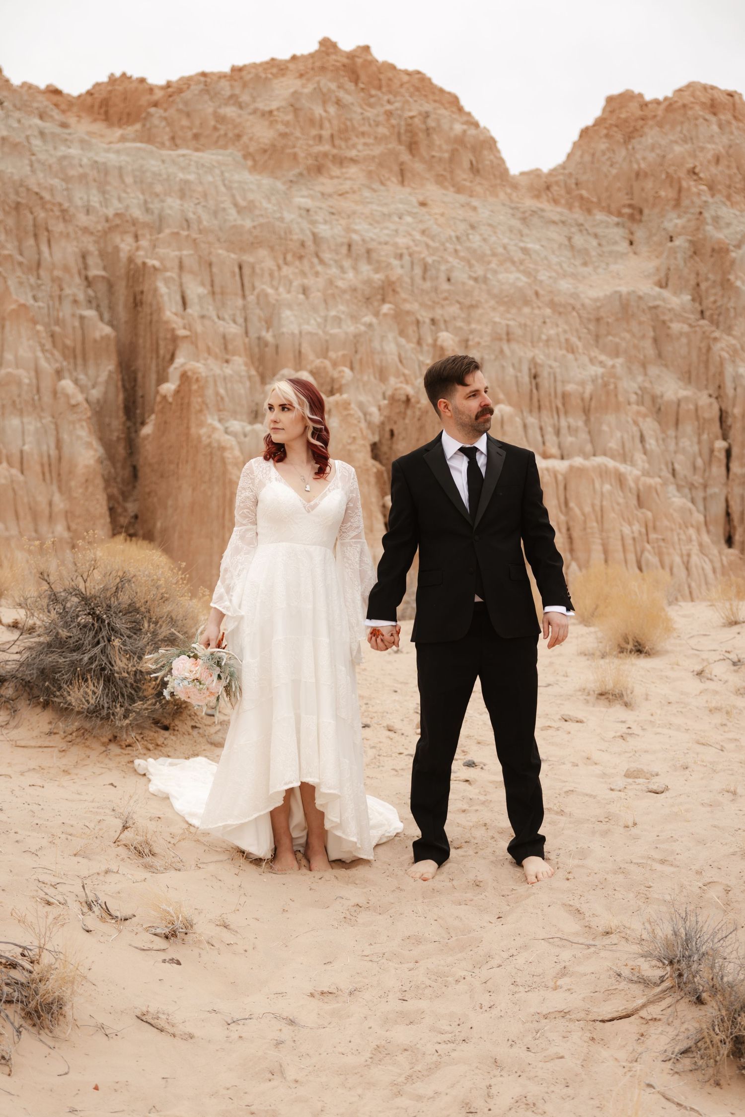 Bride in lace white gown and groom in black suit hold hands barefoot in desert sand.