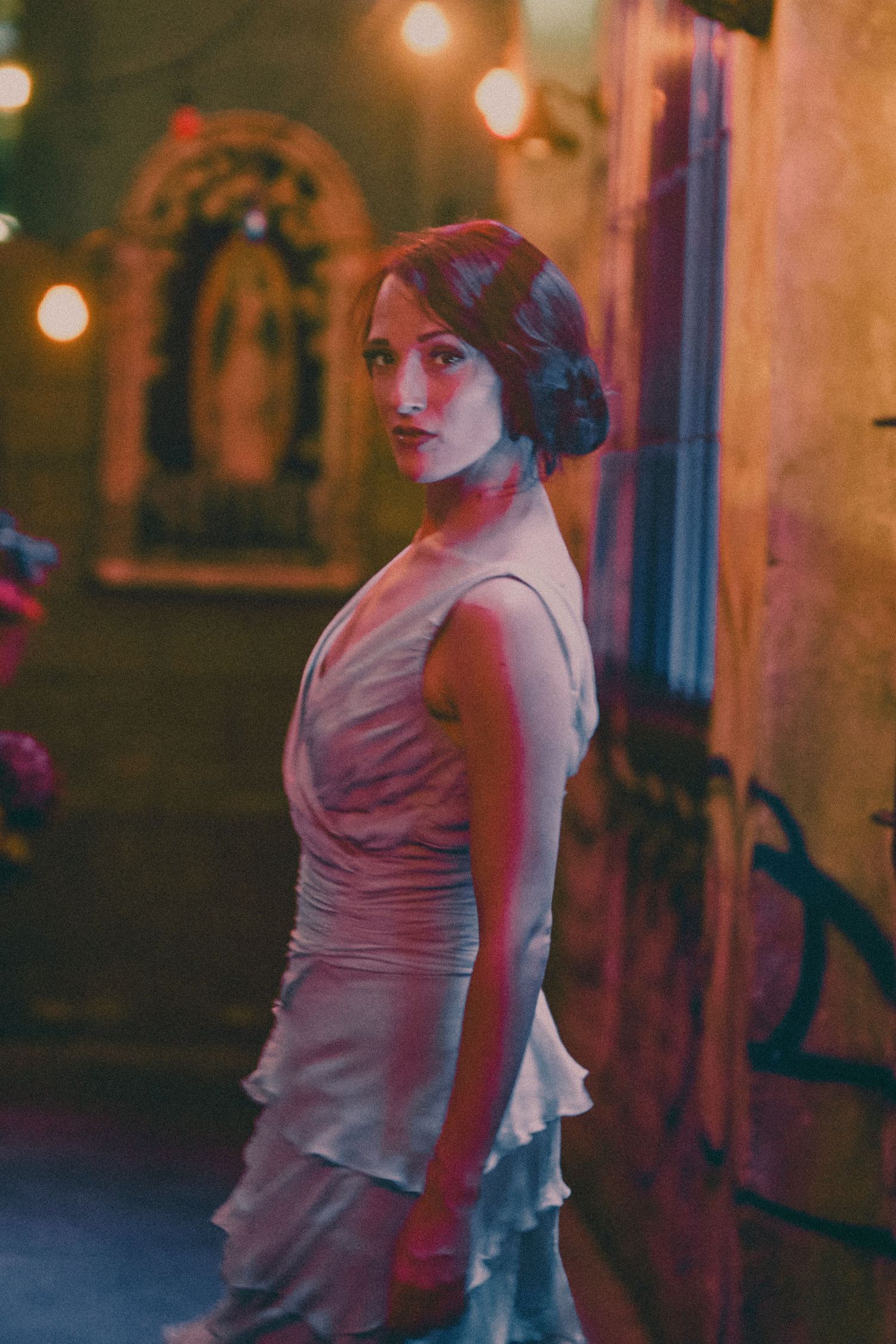 Woman in white ruffled dress poses under warm string lights in a moody, graffiti-adorned venue at night.