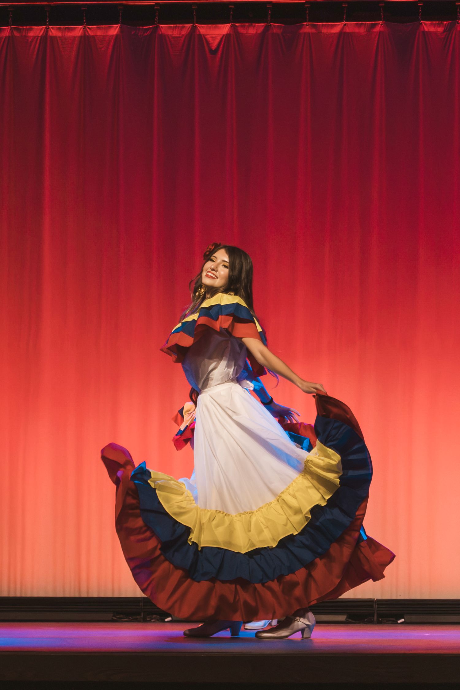 Woman in colorful traditional Colombian dress performing folk dance on stage with red curtain backdrop.