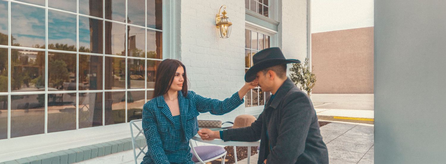 A couple holding hands at an outdoor cafe, woman in teal blazer and man in black hat, sharing a romantic moment.