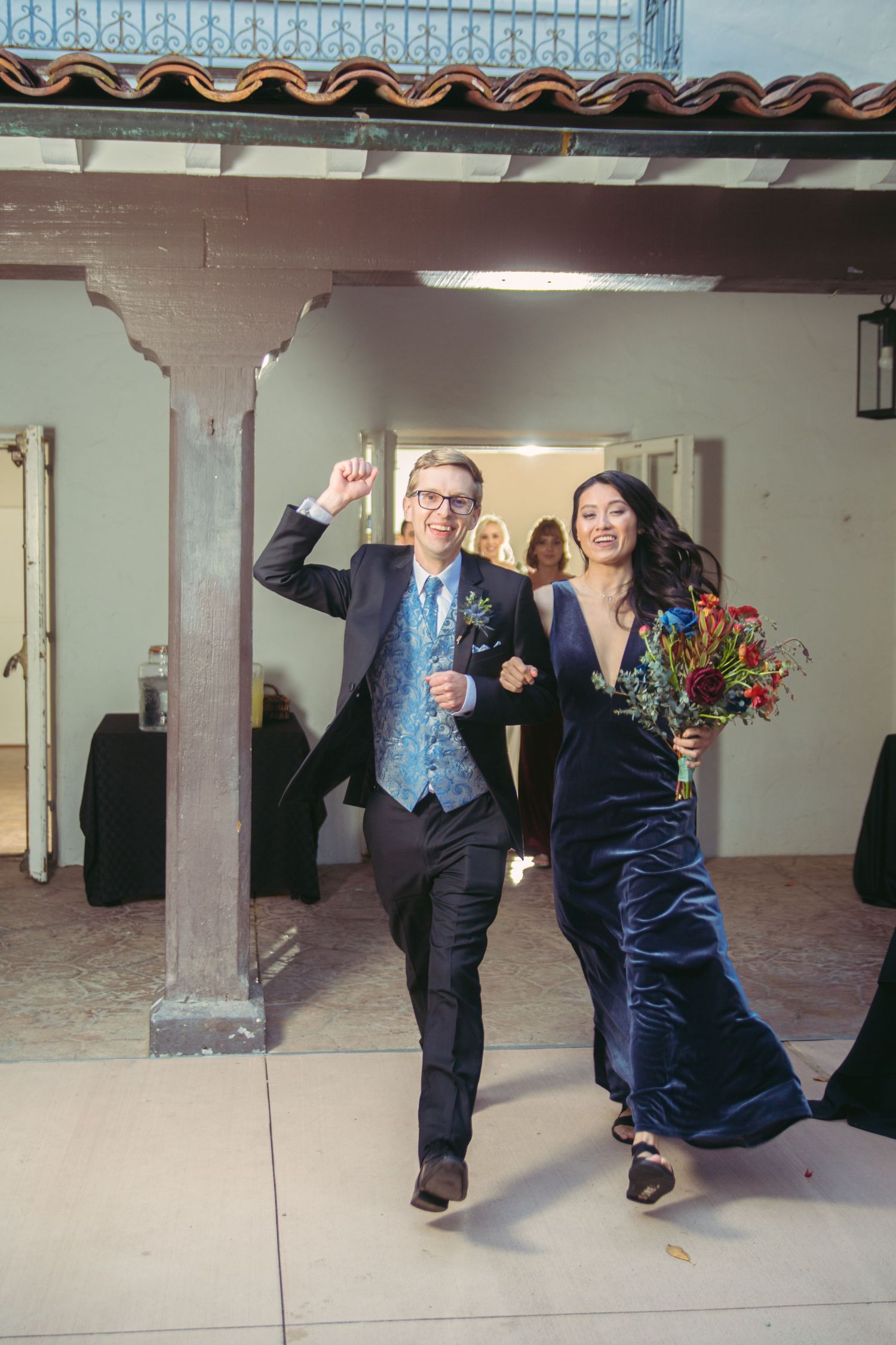Couple in navy velvet dress and blue vest enthusiastically entering a wedding reception with a floral bouquet.