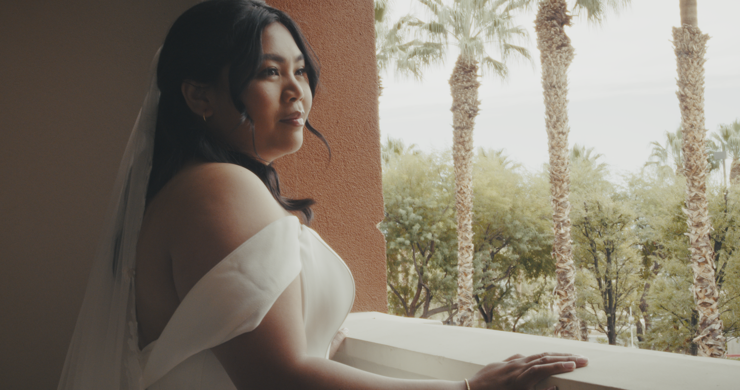 A bride in a white gown and veil gazes at palm trees from a balcony.