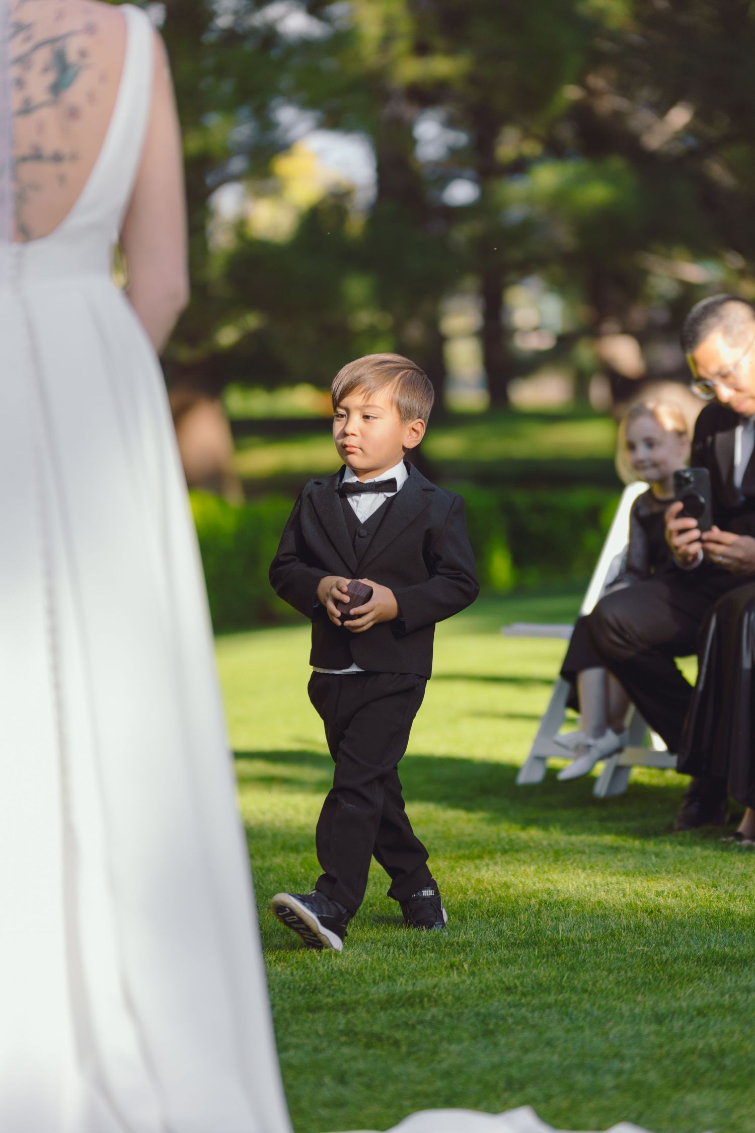 Young ring bearer in black tuxedo walking down the aisle at outdoor wedding.
