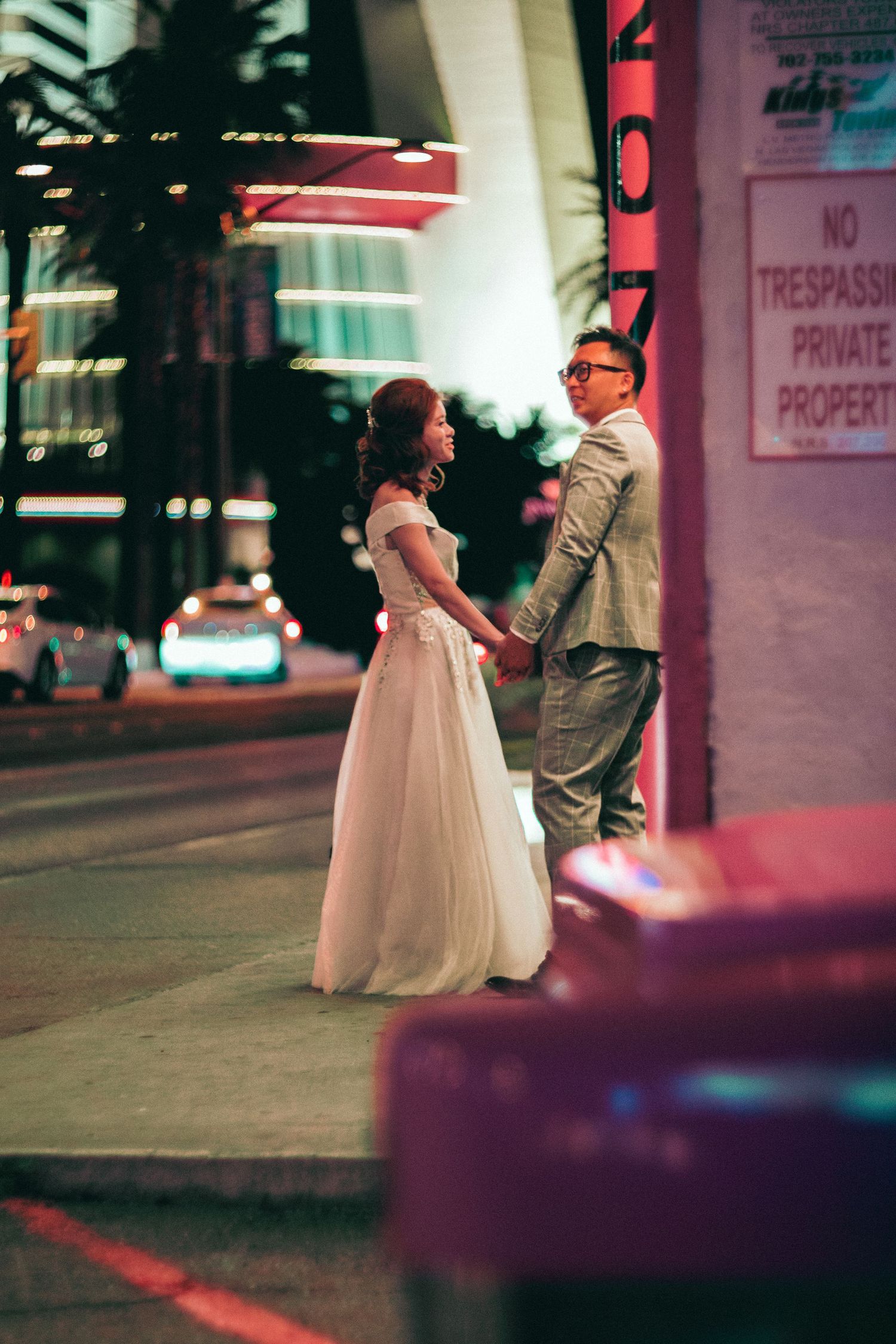 Couple in wedding attire holding hands on a city street at night with neon lights glowing in the background.