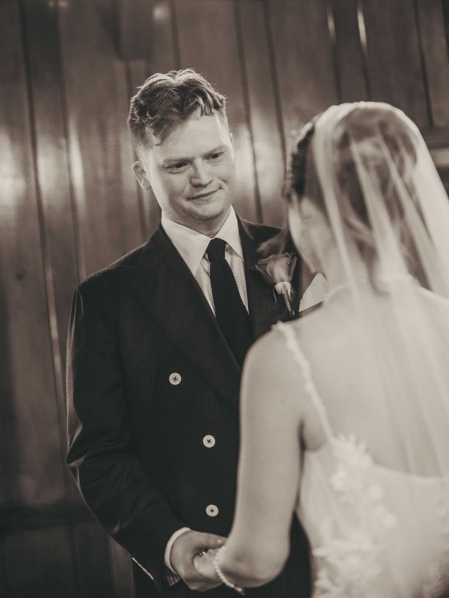 Groom smiling tenderly at bride during wedding ceremony, black and white photo capturing an emotional exchange of vows.