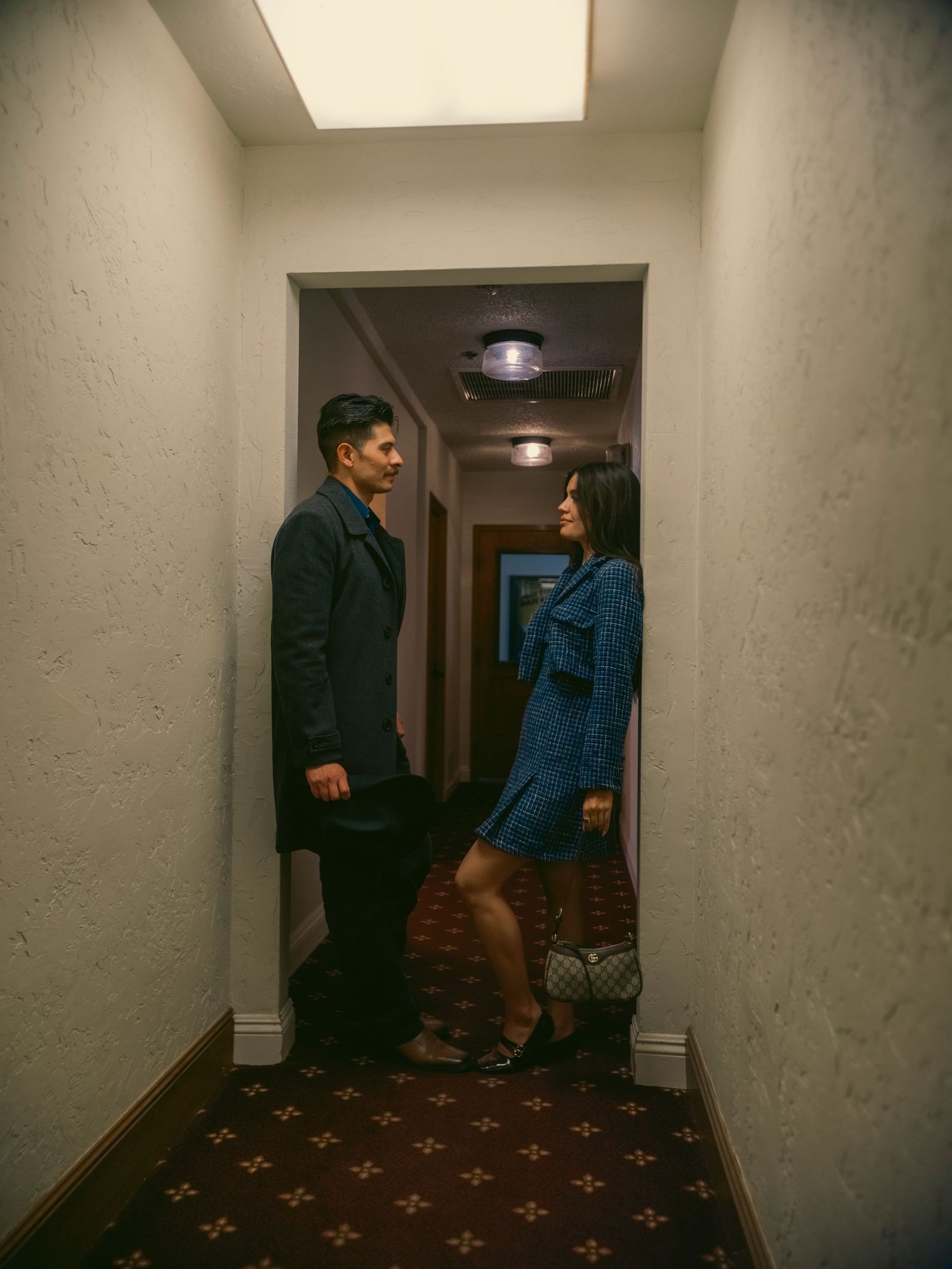 A man and woman in elegant attire face each other in a dimly lit hotel corridor with patterned carpet.