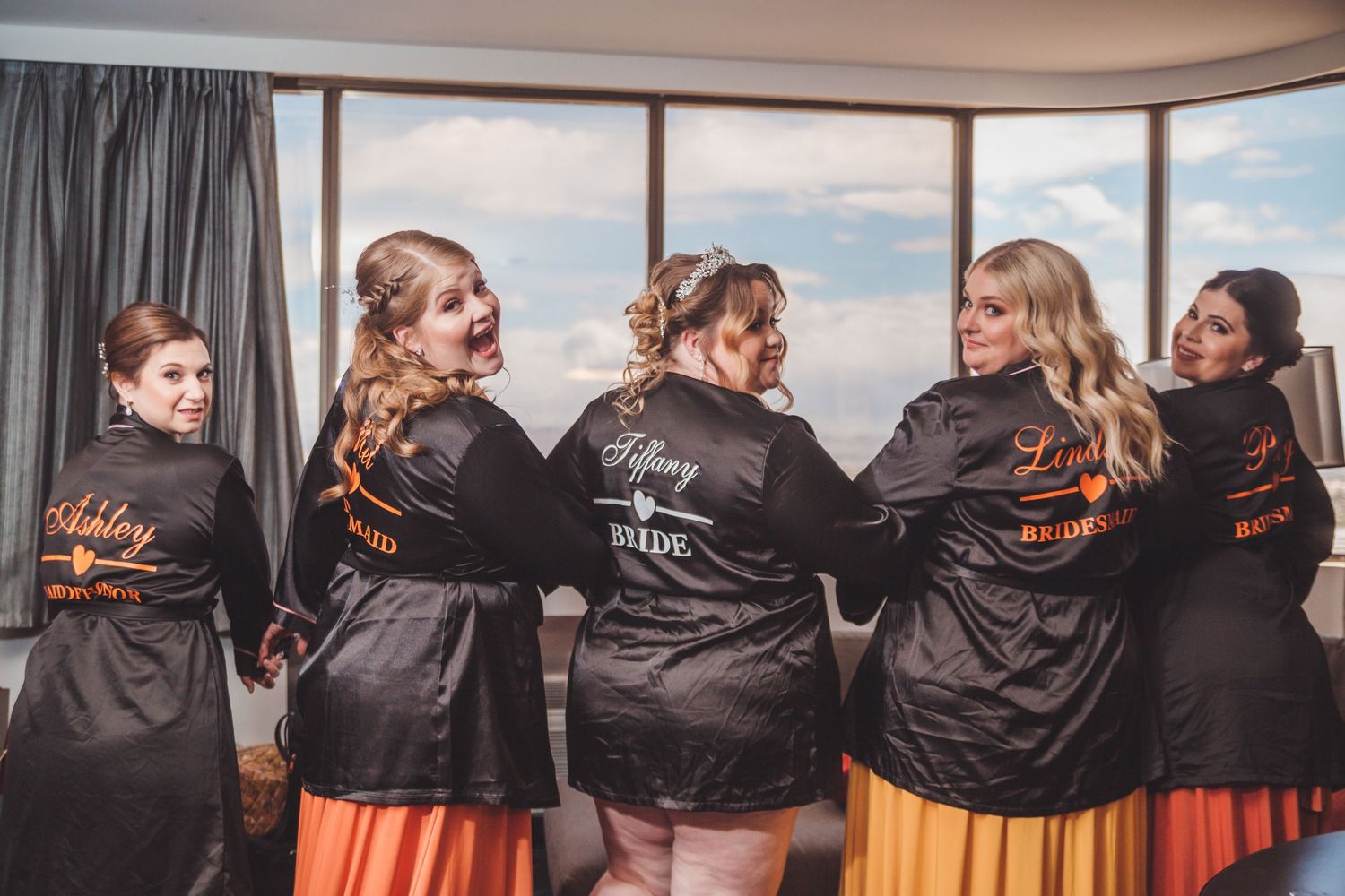 Bridesmaids wearing personalized black satin robes with orange accents pose together laughing before a wedding ceremony.