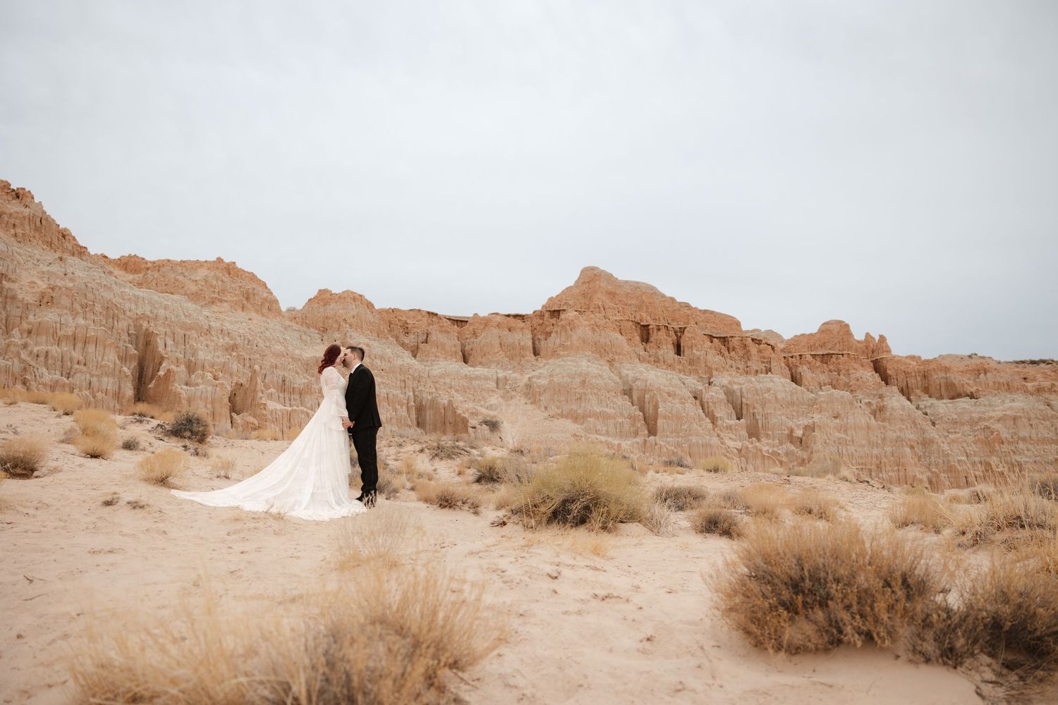 Bride and groom stand facing each other in vast desert landscape with dramatic sandstone cliffs backdrop.