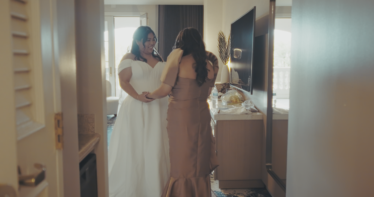 A bride in a white gown gets assisted by a woman in a bronze dress in a hotel room.