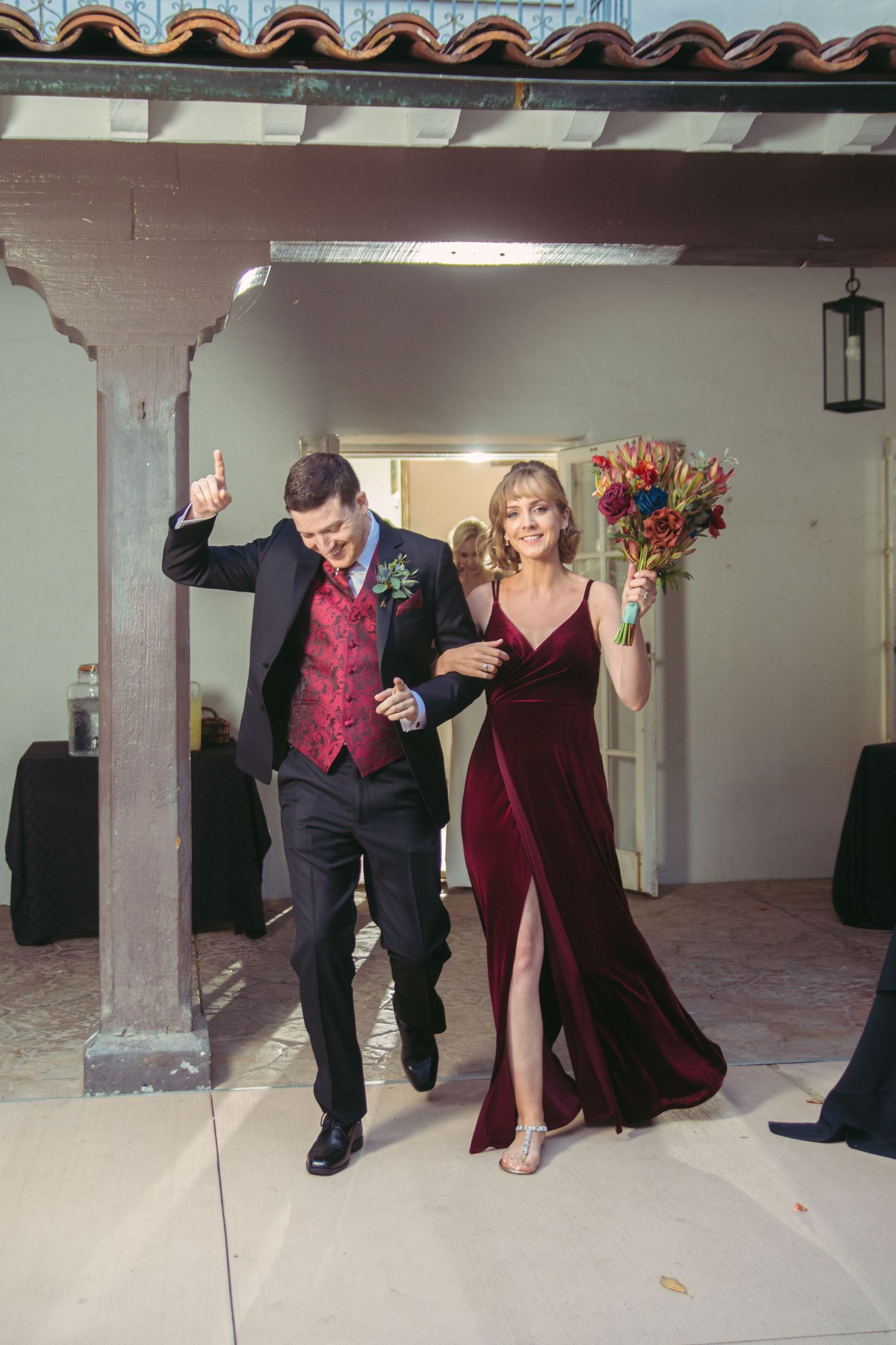 Couple in burgundy velvet gown and maroon vest making a celebratory grand entrance at a wedding reception.