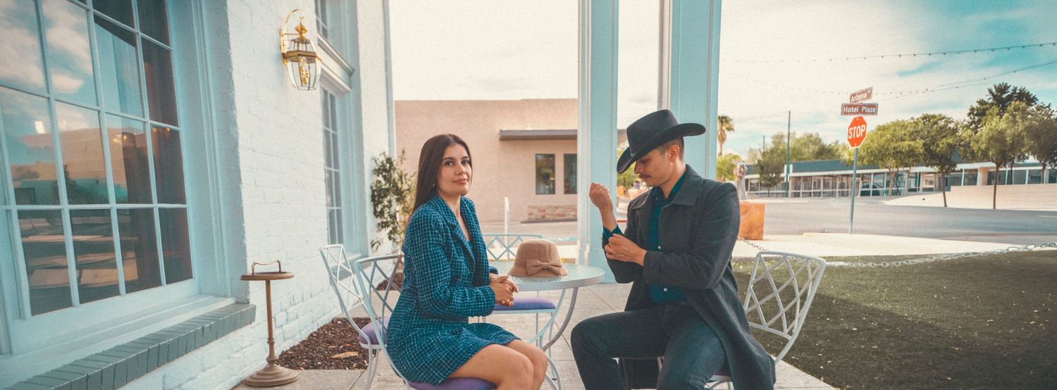A stylish couple sits at an outdoor cafe table, the woman in teal and man in a black hat, enjoying drinks on a sunny day.