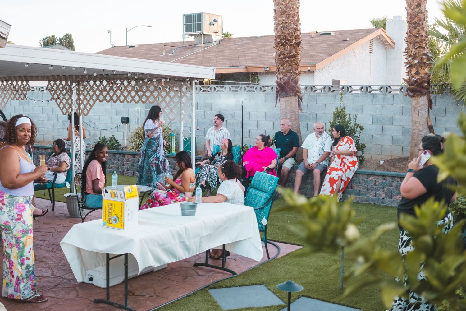 Outdoor backyard gathering with diverse group of people socializing around tables under a pergola on a sunny day.