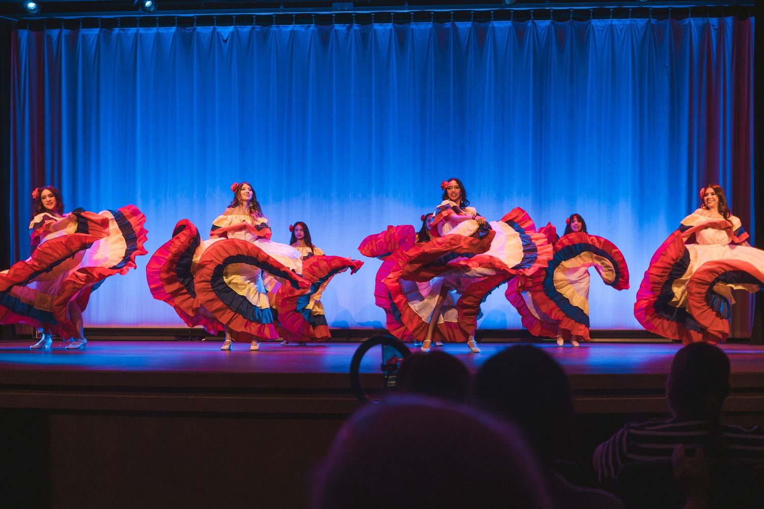 Flamenco dancers in colorful swirling dresses perform on a blue-curtained stage under warm stage lighting.