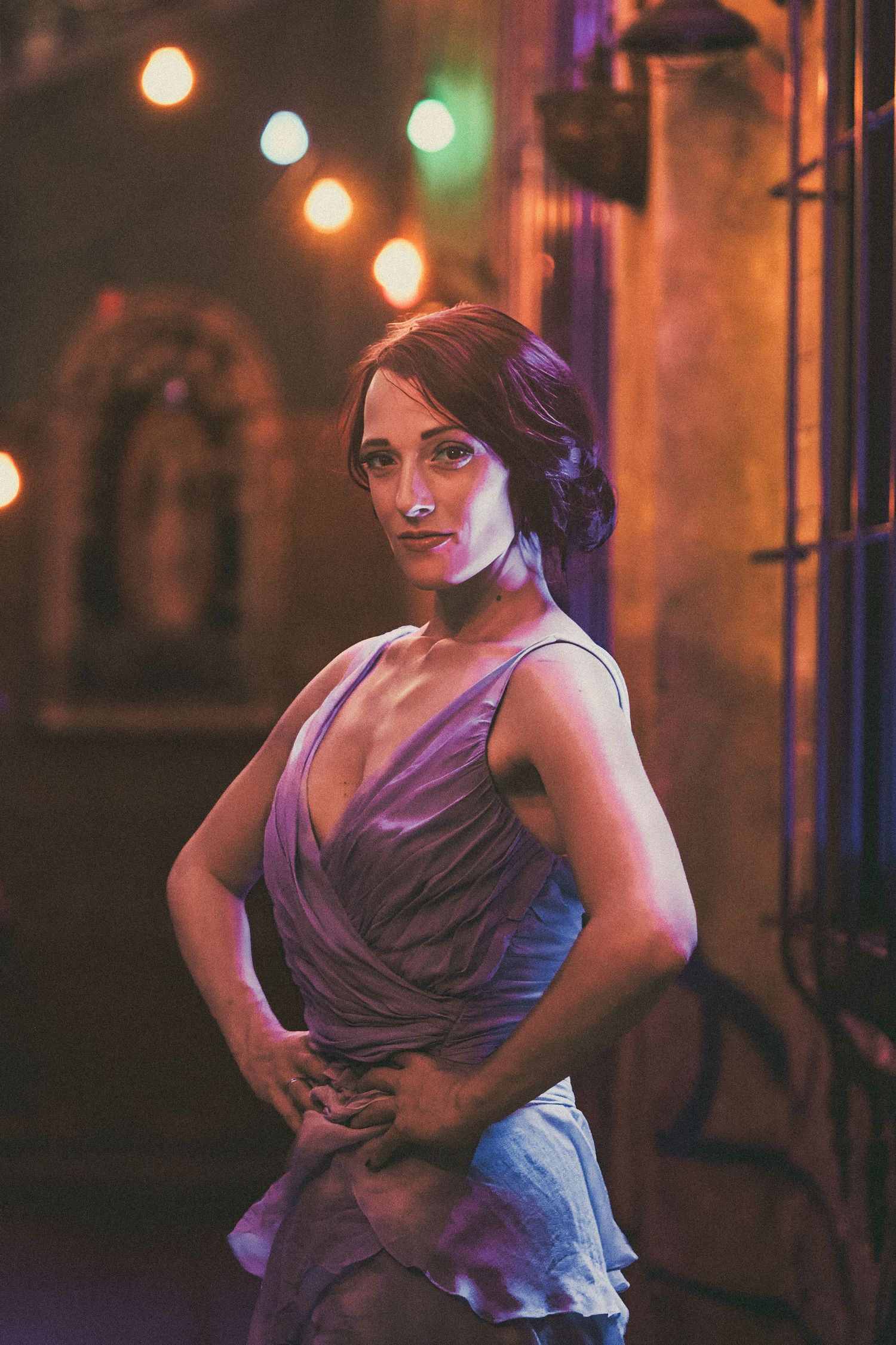 Woman in purple wrap dress poses confidently under warm string lights in a dimly lit venue.