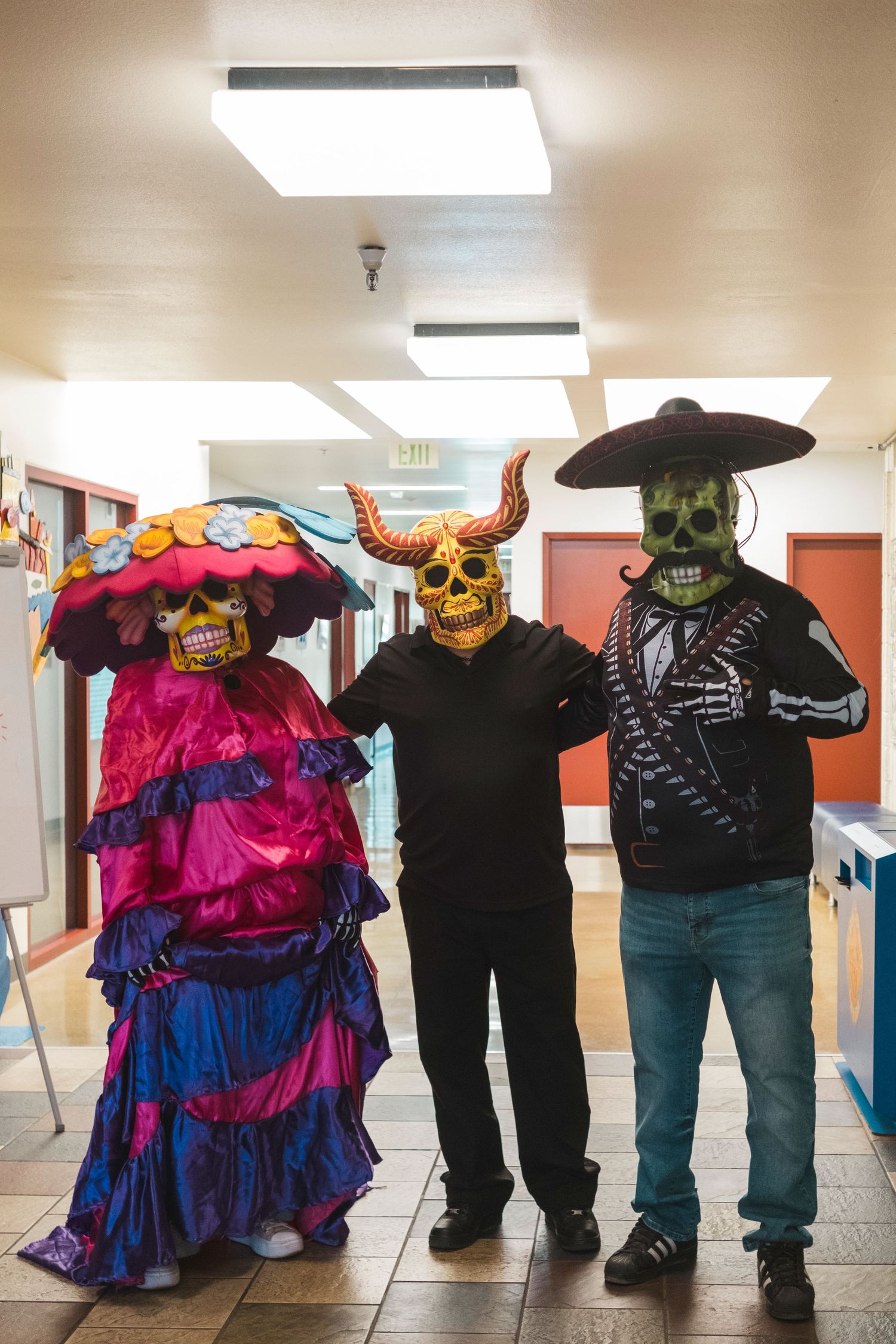 Three people in colorful Day of the Dead skull masks and costumes pose together in a hallway.