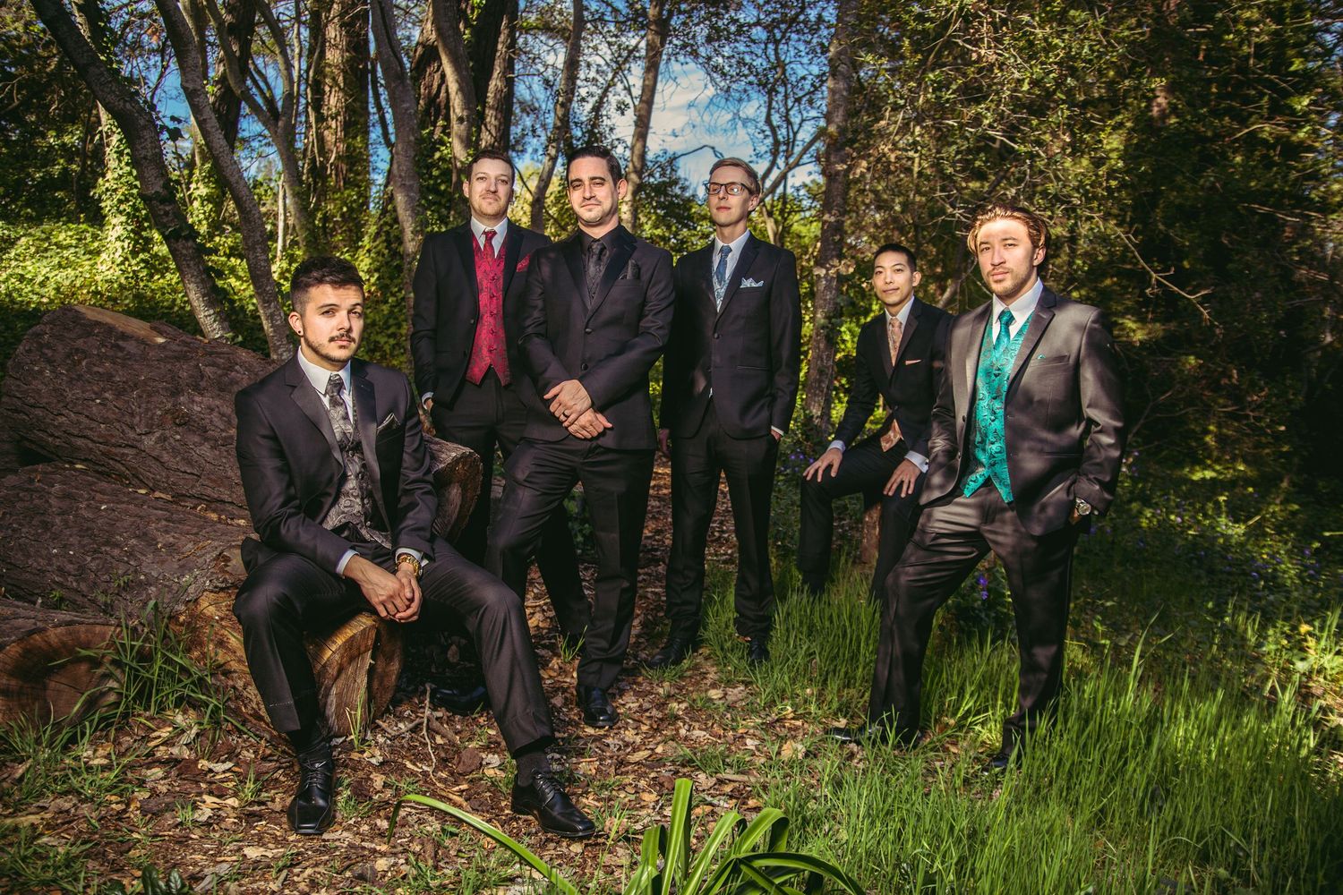 Six formally dressed groomsmen posing among green grass and trees in a natural outdoor wedding setting.