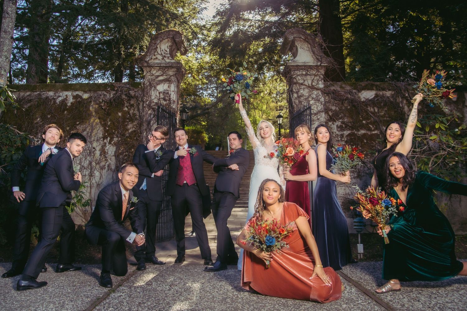 Joyful wedding party strikes fun poses at ornate stone gate, bridesmaids in colorful velvet dresses with bouquets.
