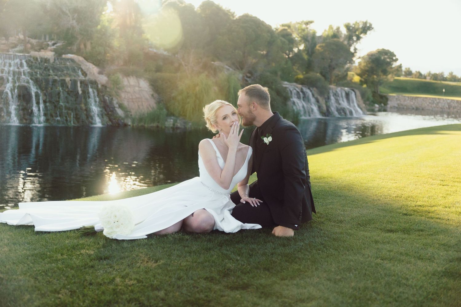 Bride and groom sitting on grass near waterfall in golden sunset light.