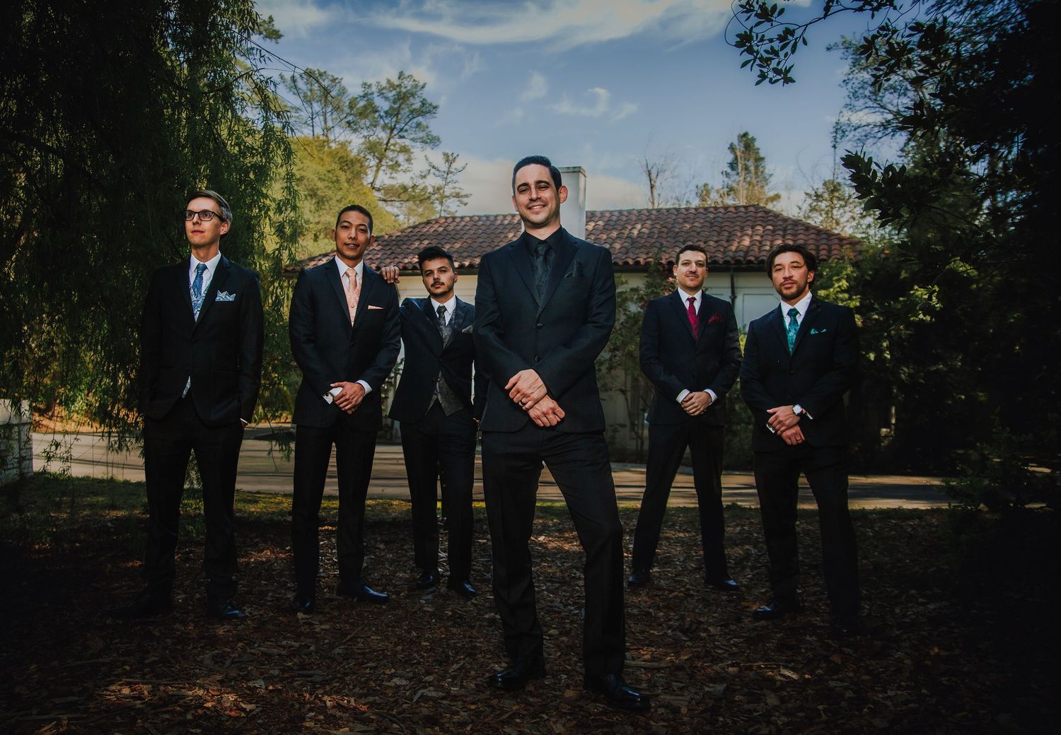 Groom stands centered with six groomsmen in black suits outdoors near a rustic building surrounded by trees.