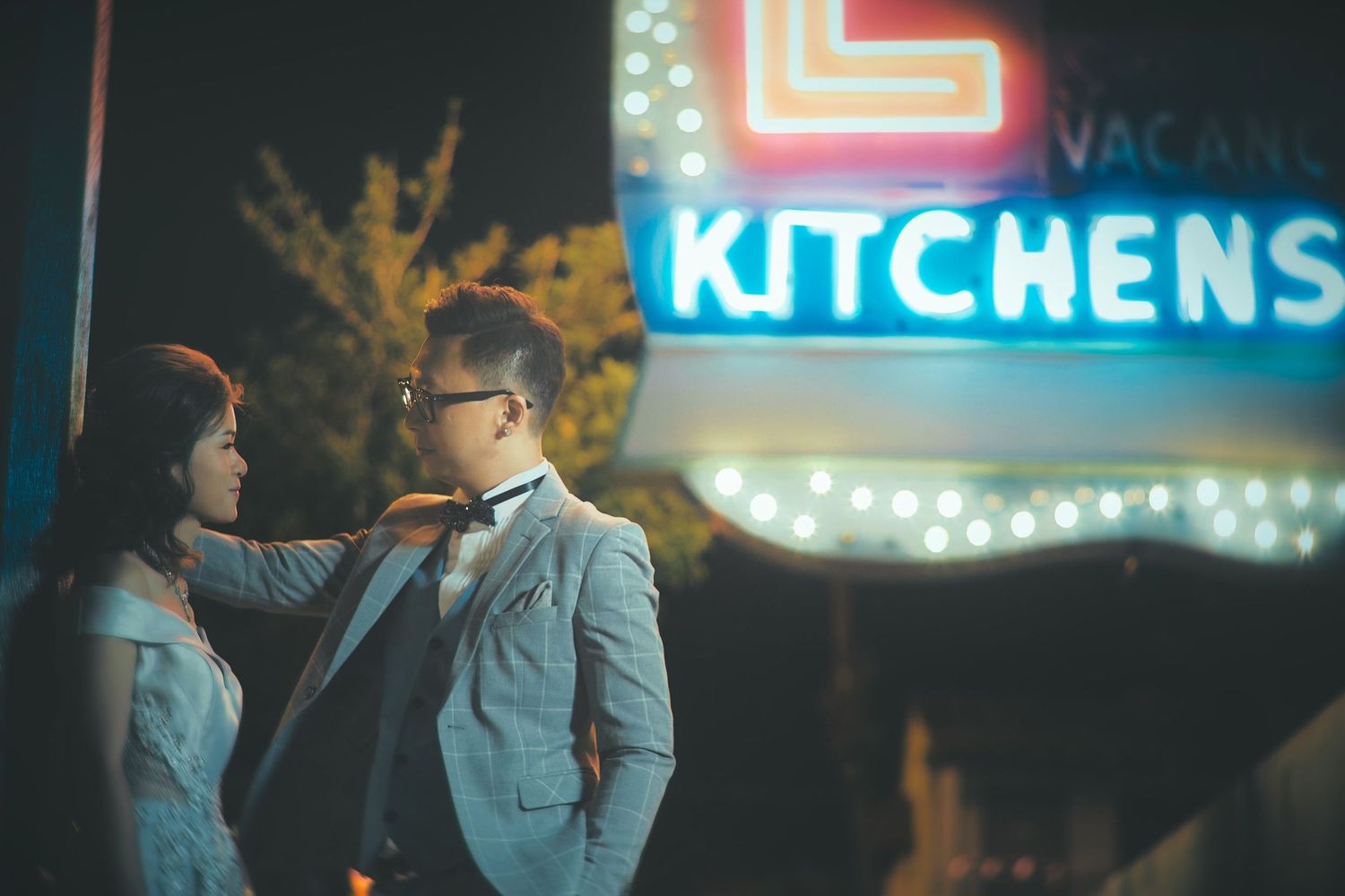 Couple poses romantically at night in front of glowing neon Kitchens sign, woman in white dress adjusting mans gray suit.