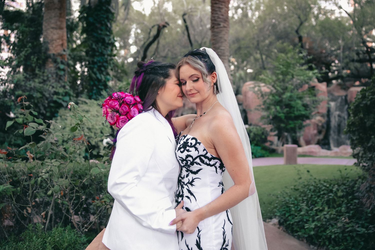 Two brides embrace and kiss in a lush garden, one wearing black and white gown with veil.