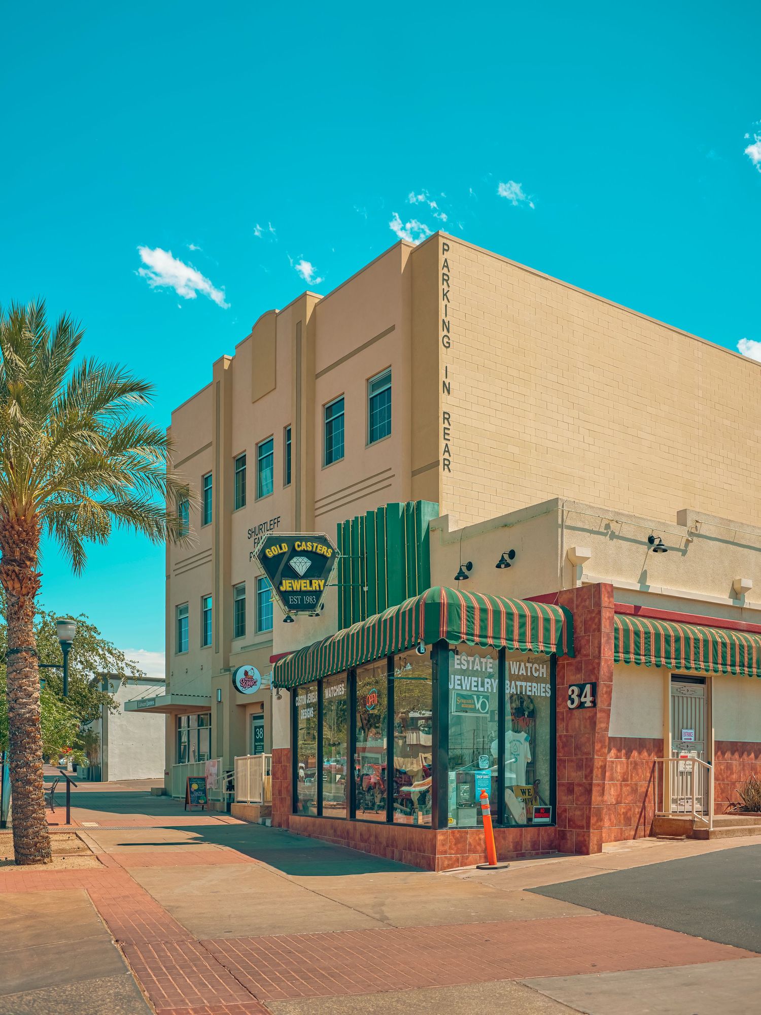 Art Deco jewelry store corner building with green awnings, palm trees, and brick sidewalk under bright blue sky.