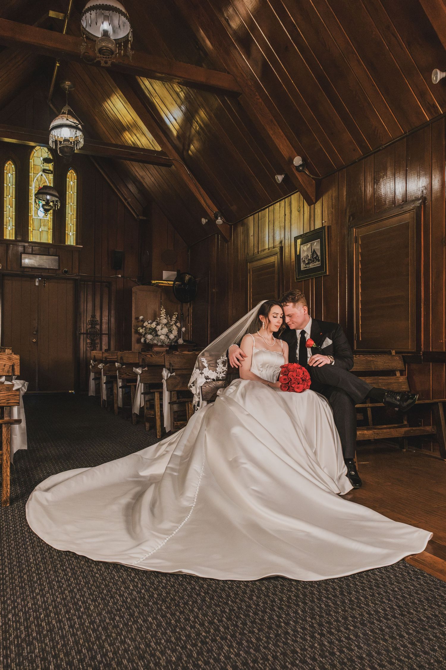 Bride and groom share a romantic moment in a wooden chapel, her white gown trailing elegantly with a red bouquet.