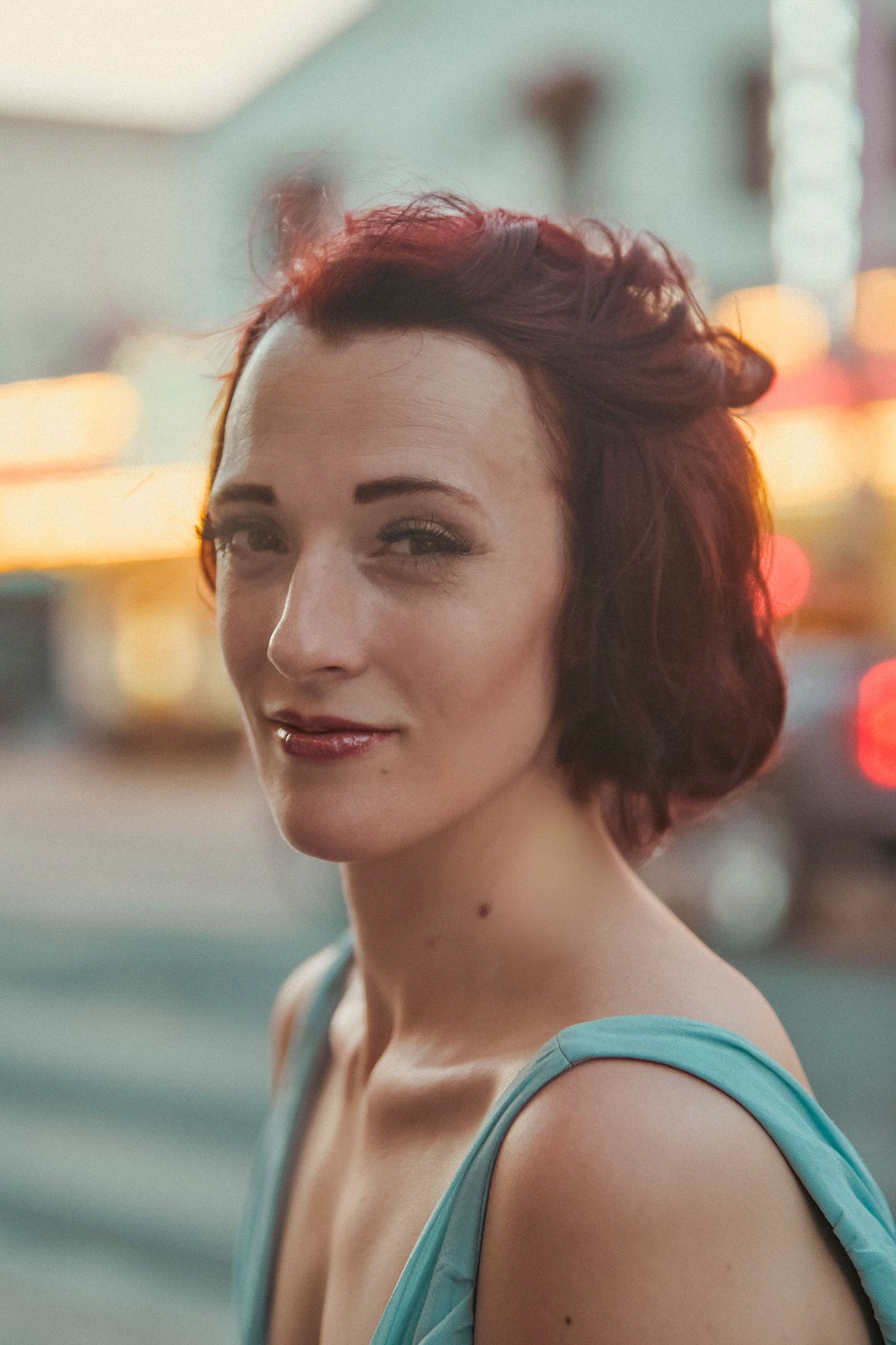 Young woman with short red hair wearing a teal dress poses outdoors at dusk with bokeh city lights behind her.