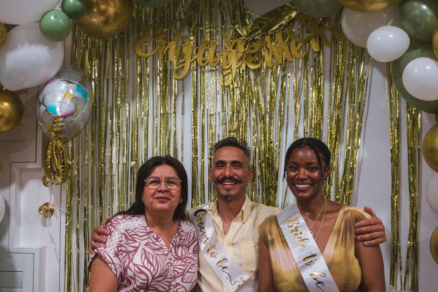 Three people posing at a festive party with gold balloon arch and shimmery fringe backdrop, two wearing celebratory sashes.