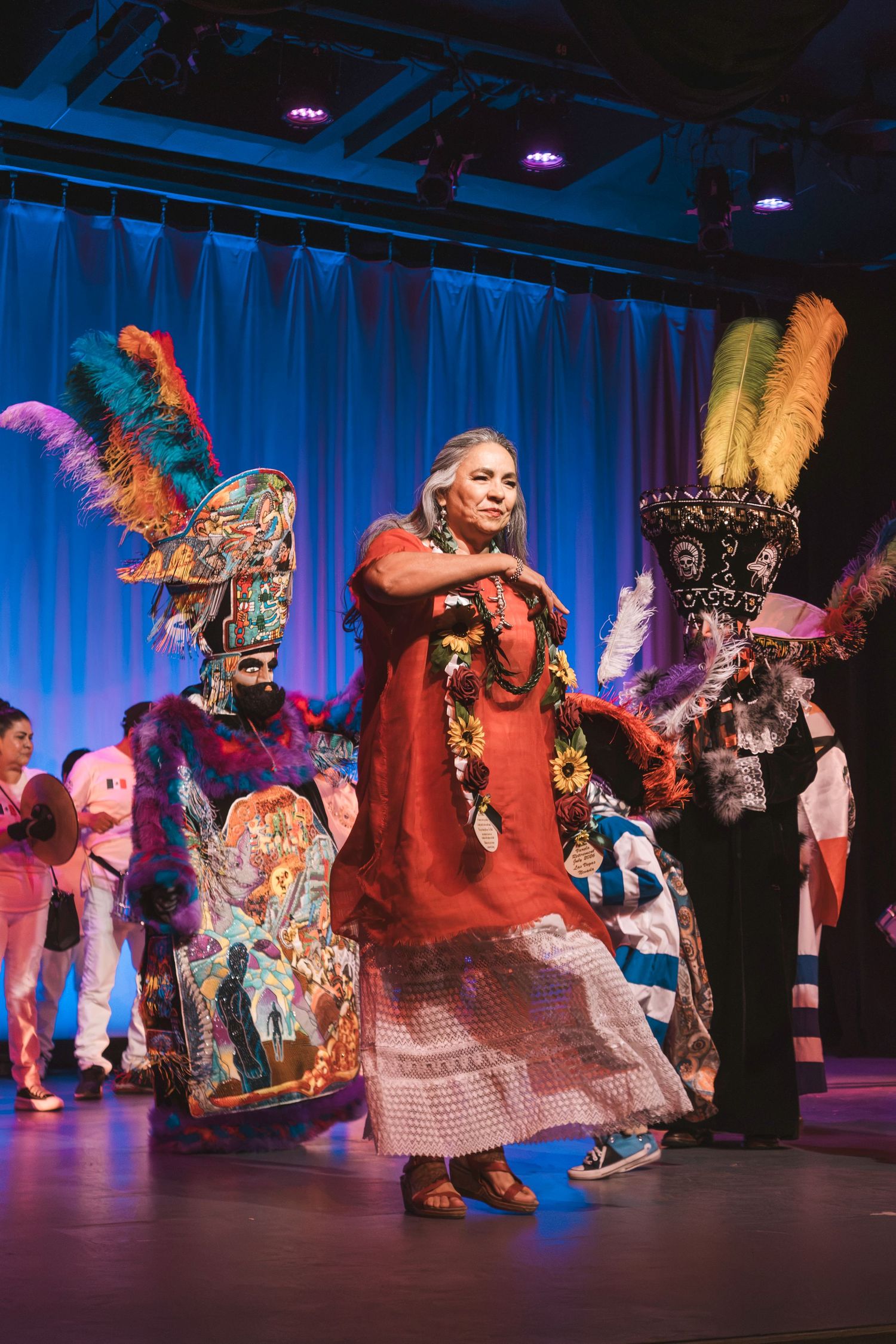 Woman in red dress dances on stage with colorful feathered headdress performers at a cultural celebration.